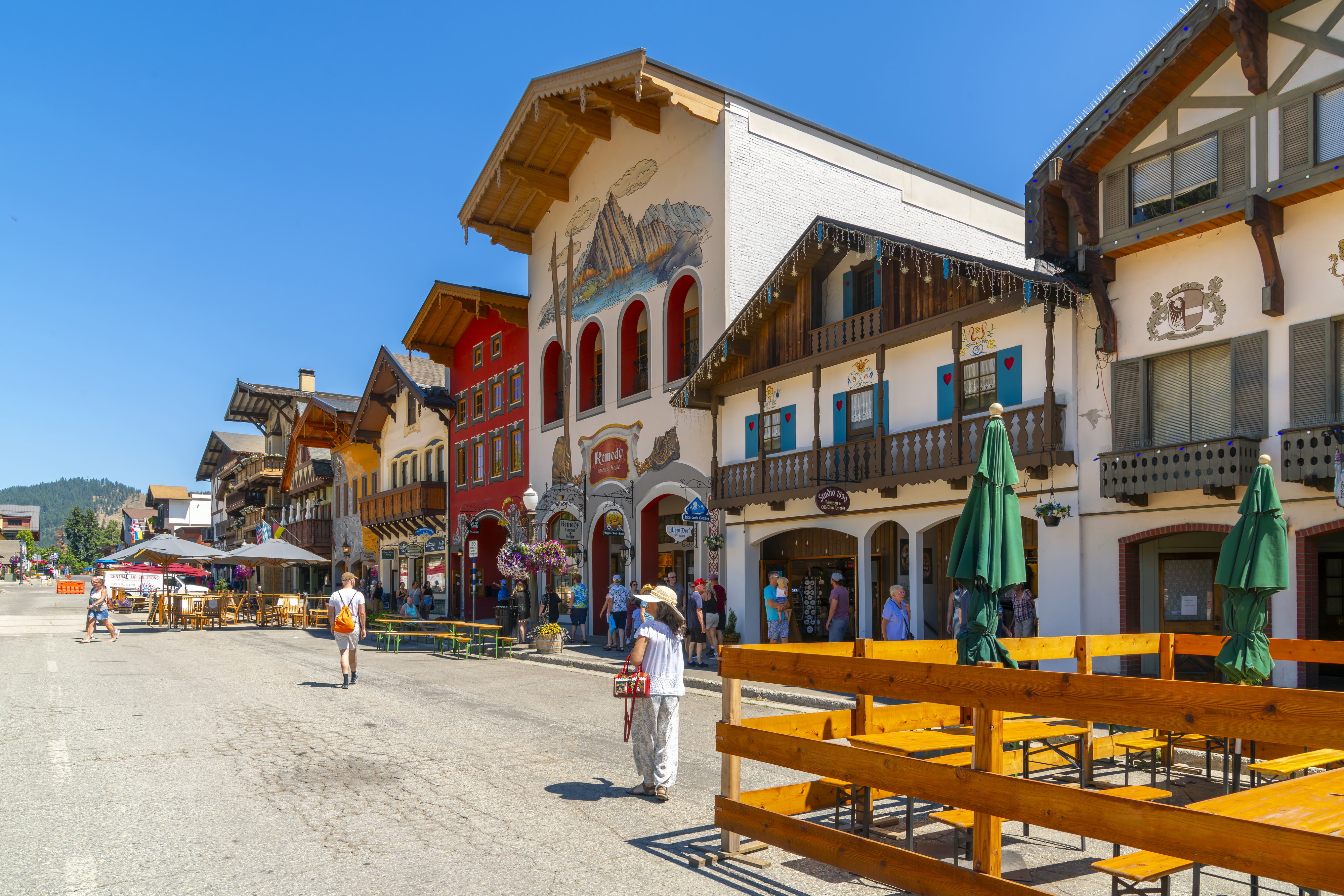 People walking and sitting by outdoor restaurants in a town with buildings designed in a Bavarian style. The sky is clear and sunny