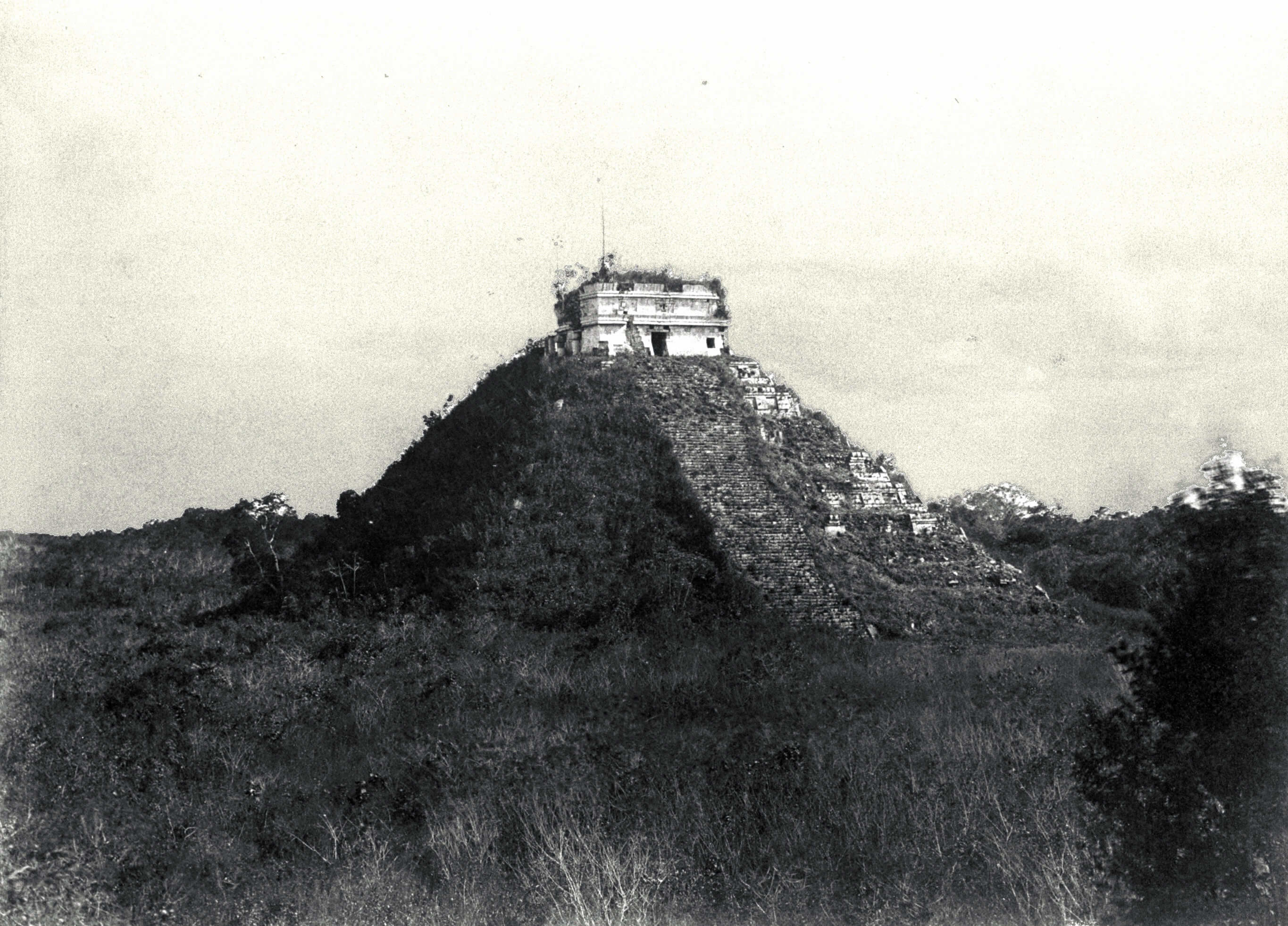 An ancient pyramid stands atop a steep hill surrounded by dense vegetation. The structure appears weathered and historic