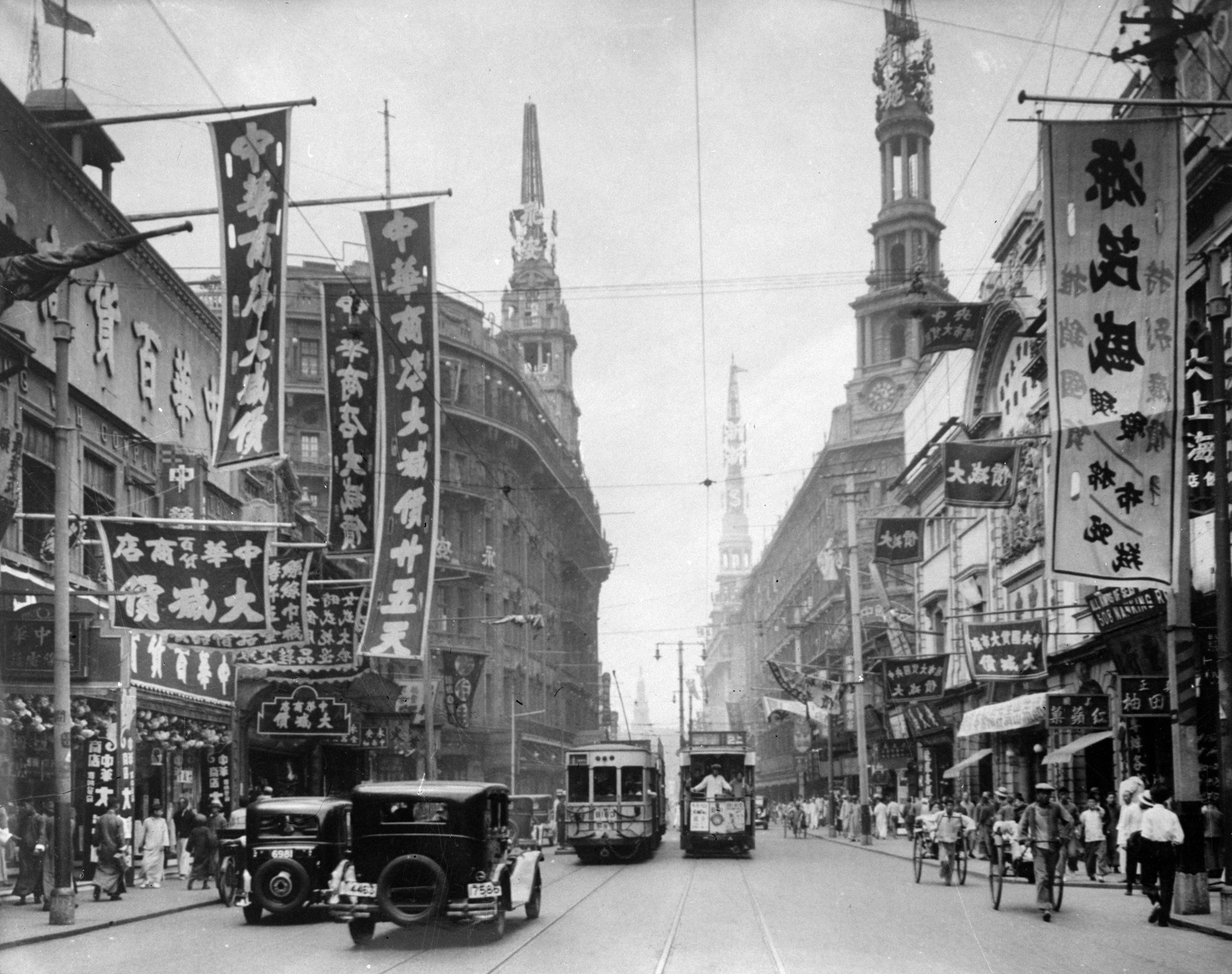 Historical photo of a busy street in Shanghai with vintage cars, trams, bicycles, and pedestrians. The street is lined with banners displaying Chinese text