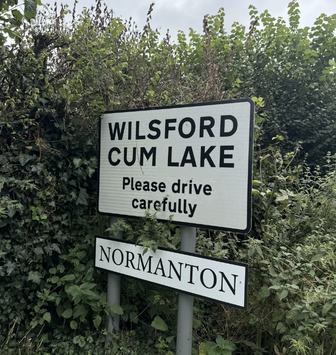 Road sign reads "Wilsford Cum Lake. Please drive carefully." A smaller sign below reads "Normanton." The signs are surrounded by greenery
