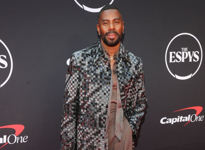 Colman Domingo on the red carpet at The ESPYS, wearing a checkered long coat over a grey pantsuit, holding sunglasses in his left hand