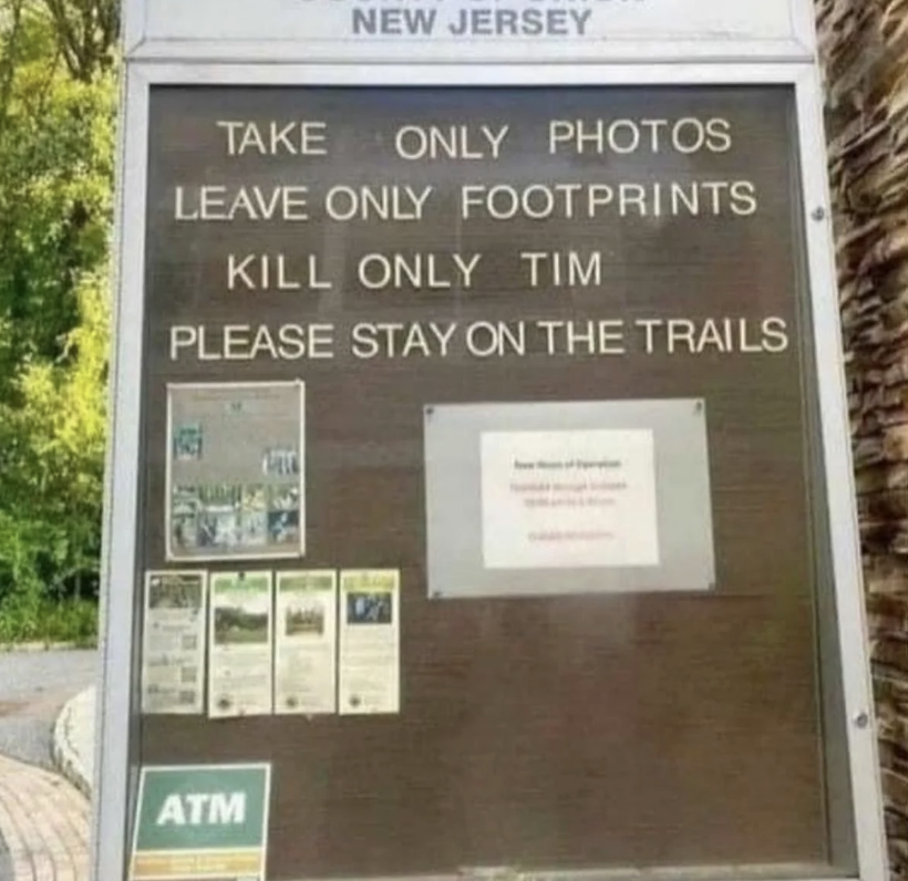 Notice board in a park with the text: "Take only photos, leave only footprints, kill only Tim, please stay on the trails." The word "Tim" appears to be an error