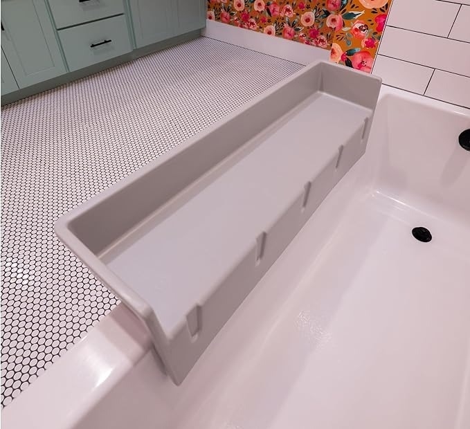 A white bathtub with a detachable gray tray covering one edge, placed against a hexagonal-tiled floor and floral-patterned wall