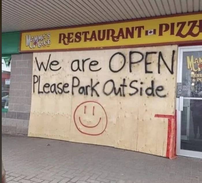 A restaurant with a boarded-up front has a spray-painted sign saying, "We are OPEN. Please Park Outside" with a smiling face drawn below the text