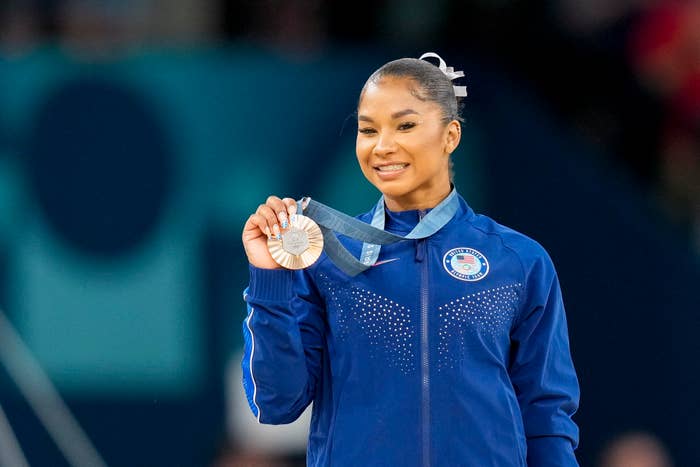 Gymnast Jordan Chiles smiles and holds up her silver medal while wearing a tracksuit adorned with rhinestones