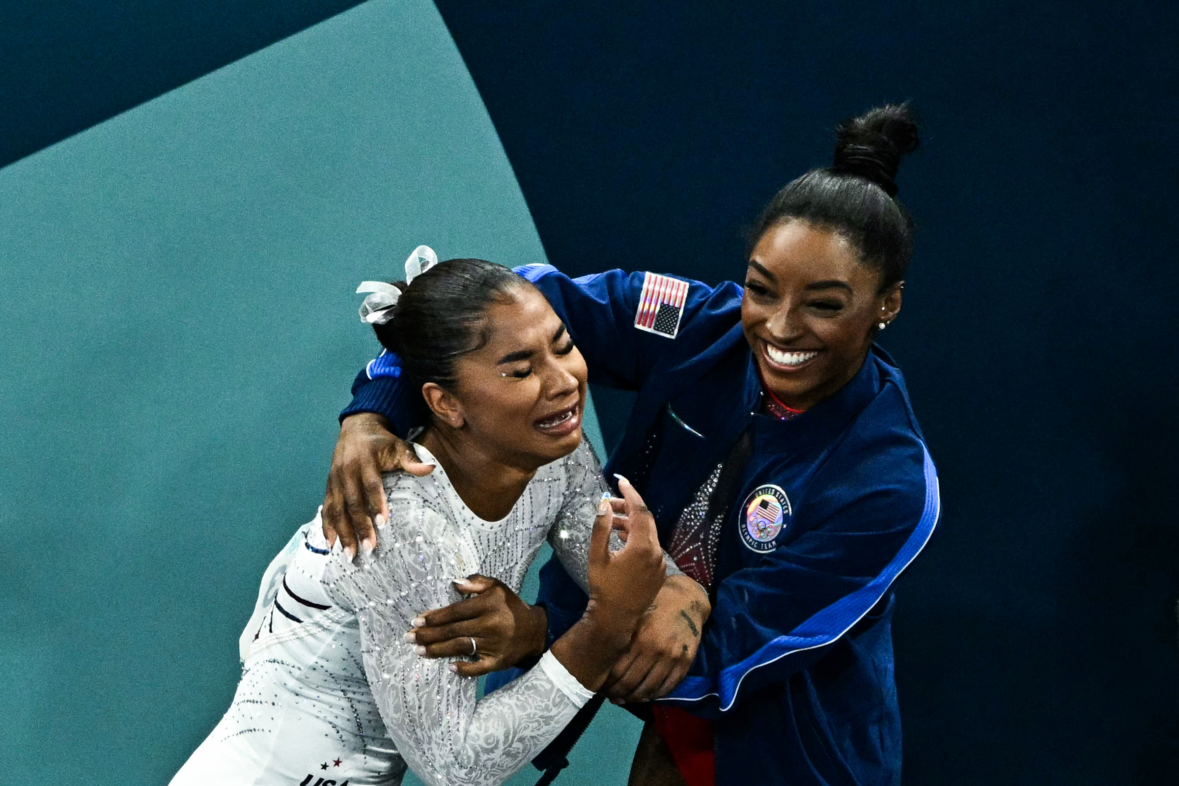 Athletes Jordan Chiles and Simone Biles share an emotional moment. Chiles appears tearful in a gymnastics uniform while Biles comforts her in a jacket