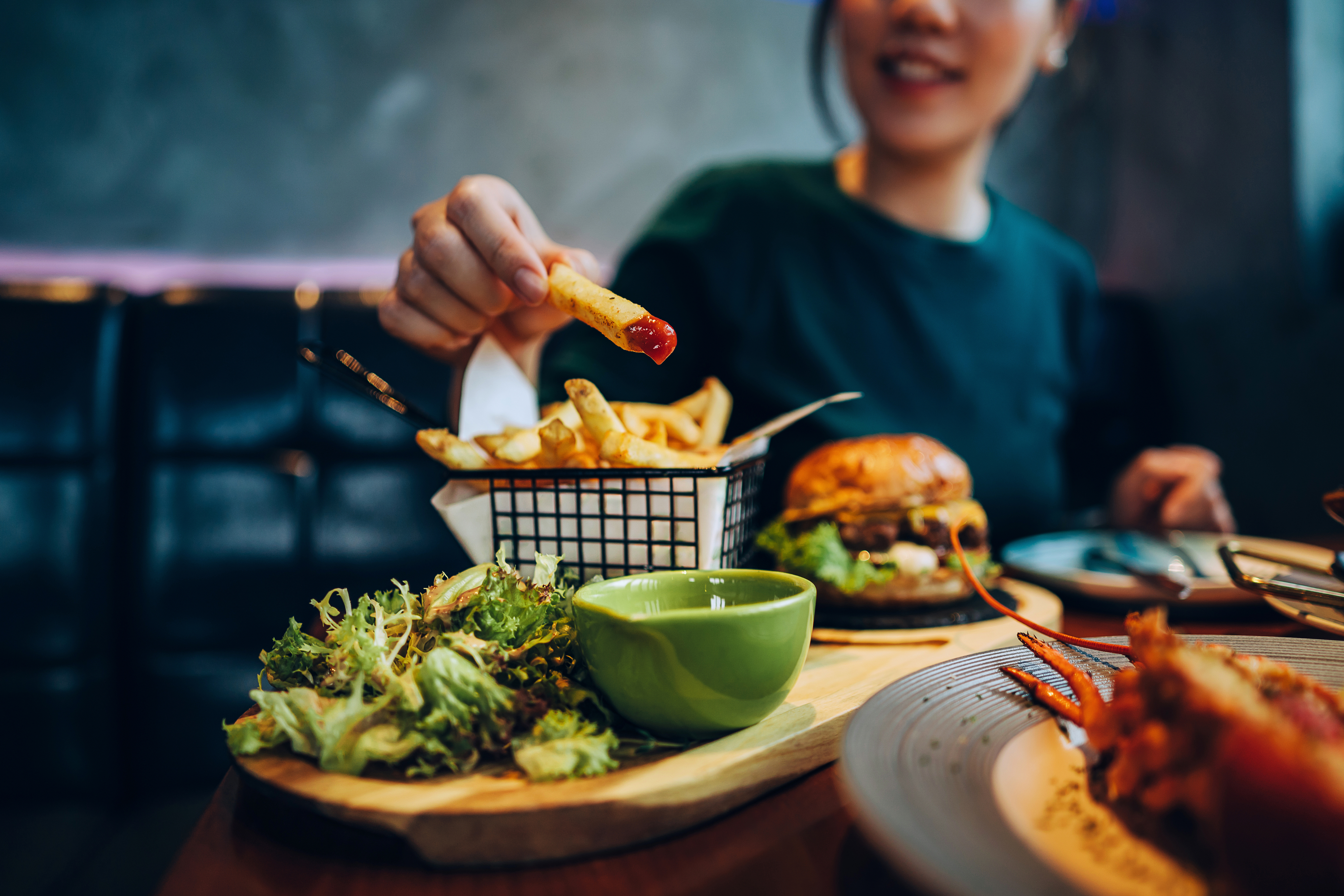 Person eating fries, burger, and salad at a restaurant table, smiling in the background