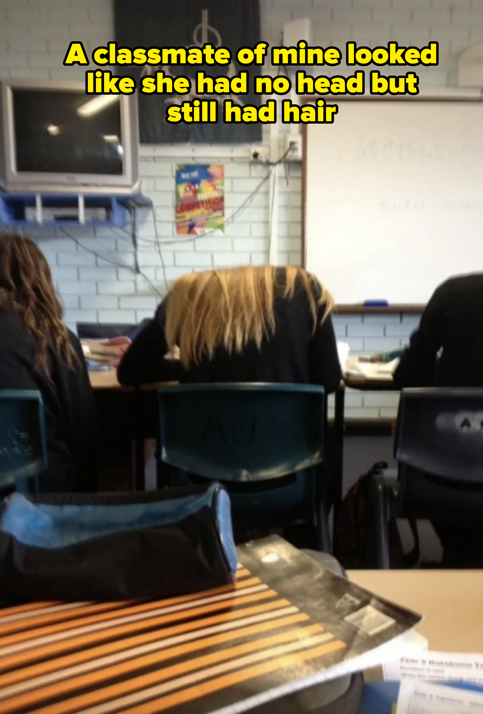 Three students sitting at desks in a classroom; one has long blonde hair. A TV is mounted on the wall above them. Whiteboard and posters are in the background