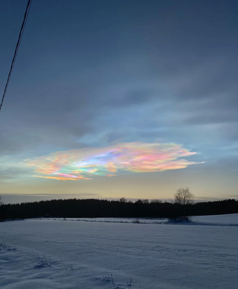 A scenic winter landscape with snow-covered fields and a distant treeline, featuring iridescent clouds in the sky