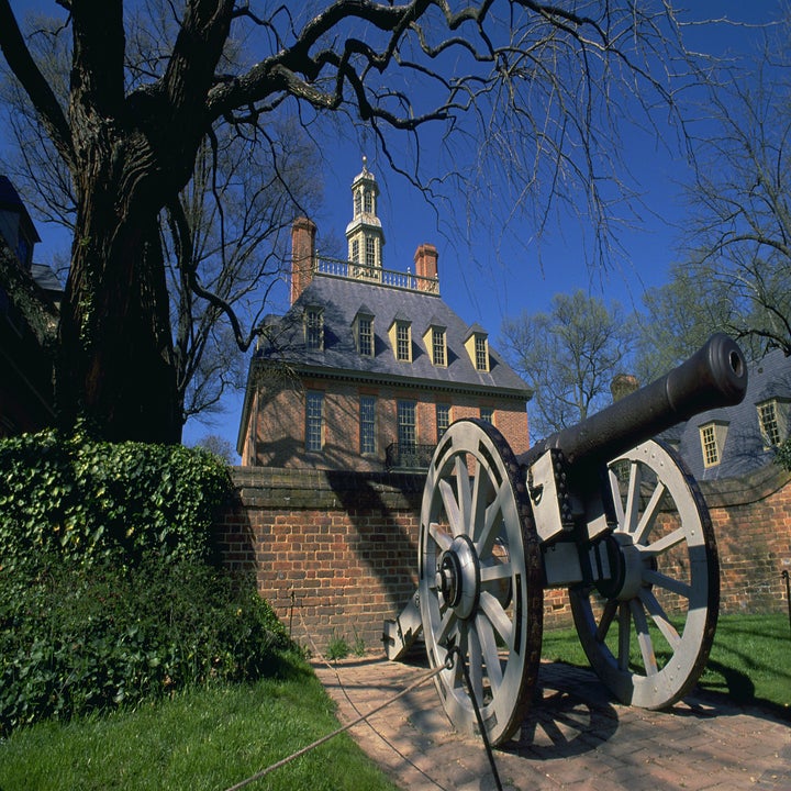 A historic cannon is displayed in front of a colonial-era brick building with a cupola and chimneys, surrounded by trees and ivy