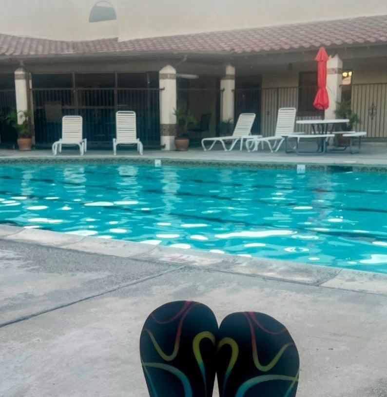 A pair of feet wearing colorful swim fins rests by a swimming pool with lounge chairs and a building in the background