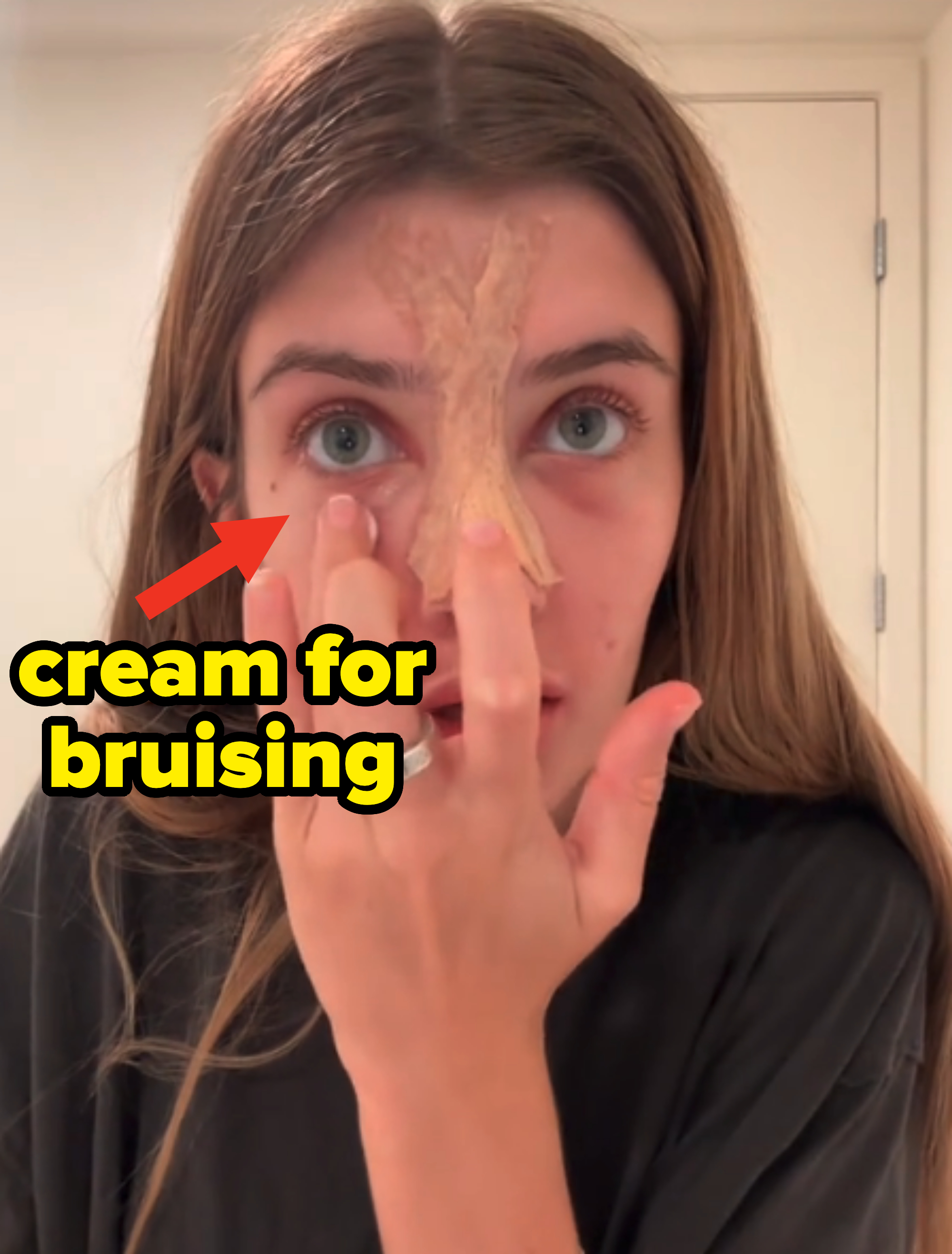 Woman in a black shirt peeling a facial mask from her forehead, partially removed. She has long hair and wears a ring on her finger, looking directly at the camera