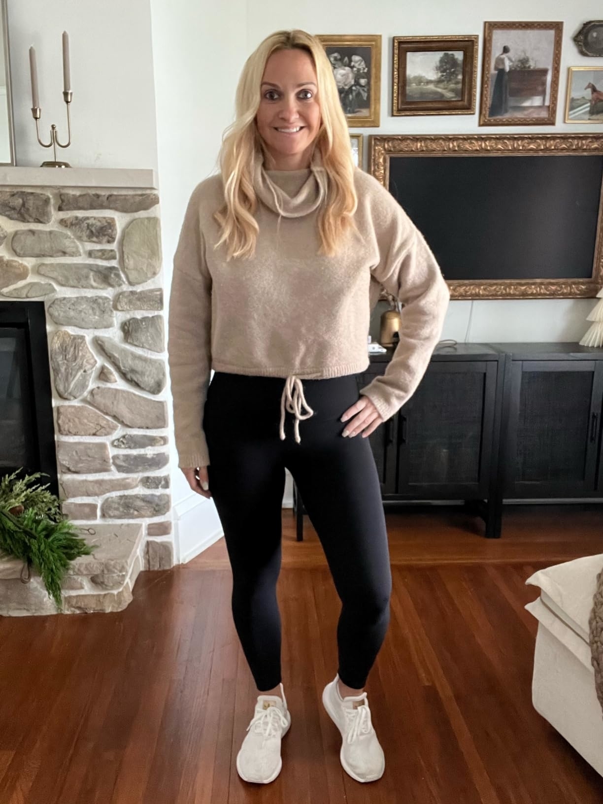 Reviewer wears a casual outfit with a beige sweater, black leggings, and white sneakers, standing in a living room with a stone fireplace and framed pictures on the wall