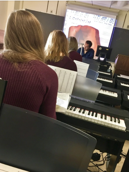 A group of music students, seated at pianos and facing forward, follows a lecture by a man at the front of the classroom while reading sheet music