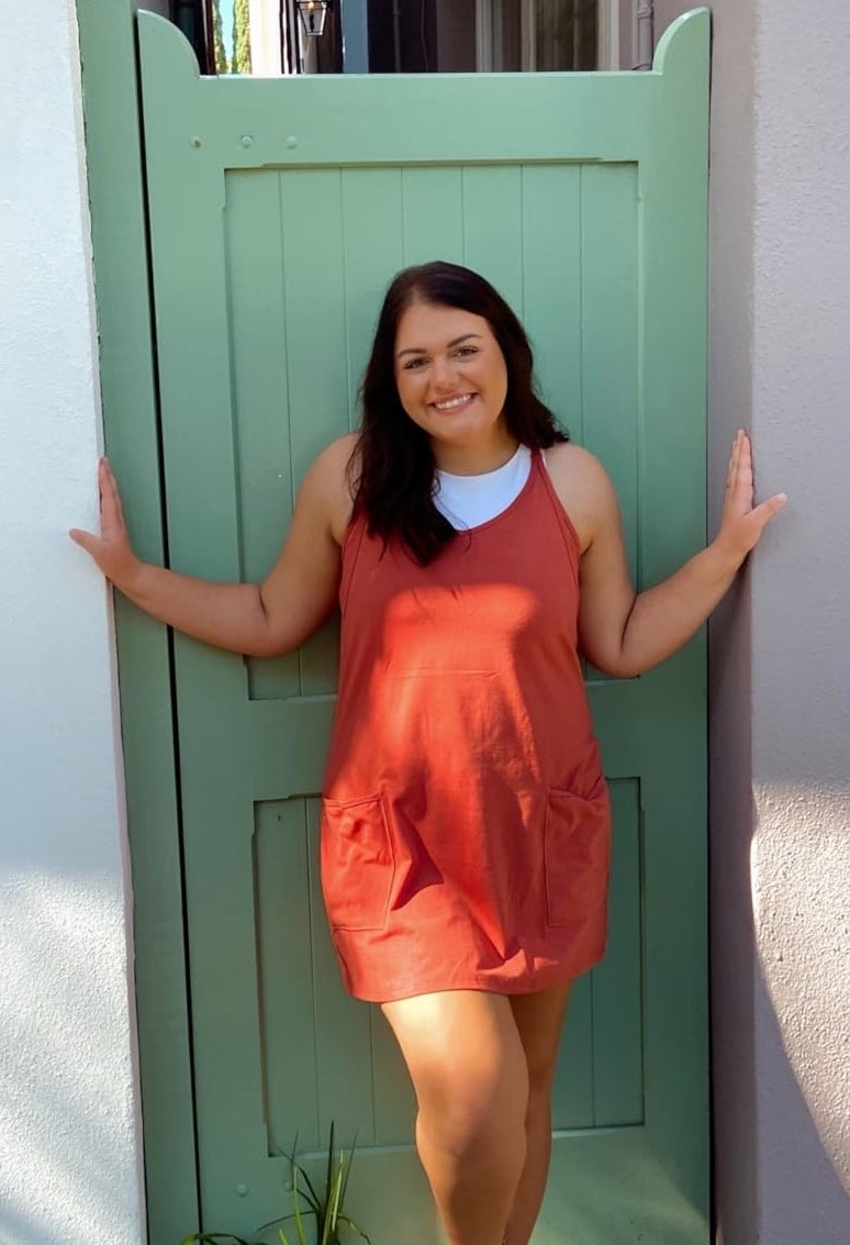 A reviewer stands in a doorway wearing a casual short dress and sneakers, smiling at the camera