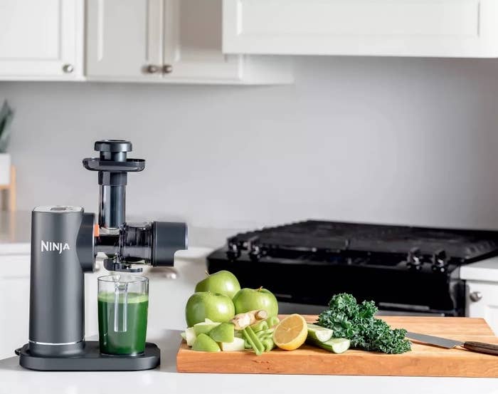 Ninja Cold Press Juicer on a kitchen counter with green juice, apples, kale, and lemon on a cutting board. Text: "Introducing the Ninja NeverClog™ Cold Press Juicer."