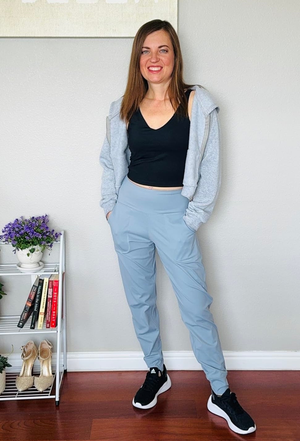 Reviewer wearing a black crop top, light gray hoodie, light gray jogger pants, and black sneakers standing next to a plant and book shelf