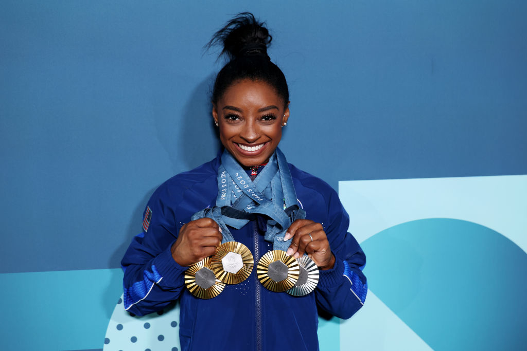 Simone Biles holding and displaying several gold medals, smiling broadly. She is wearing an athletic jacket with her hair in a high bun