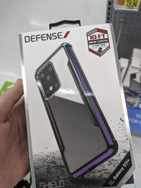 A person holds a Defense Shield phone case packaging for a Galaxy S11+, advertising 10 ft drop protection