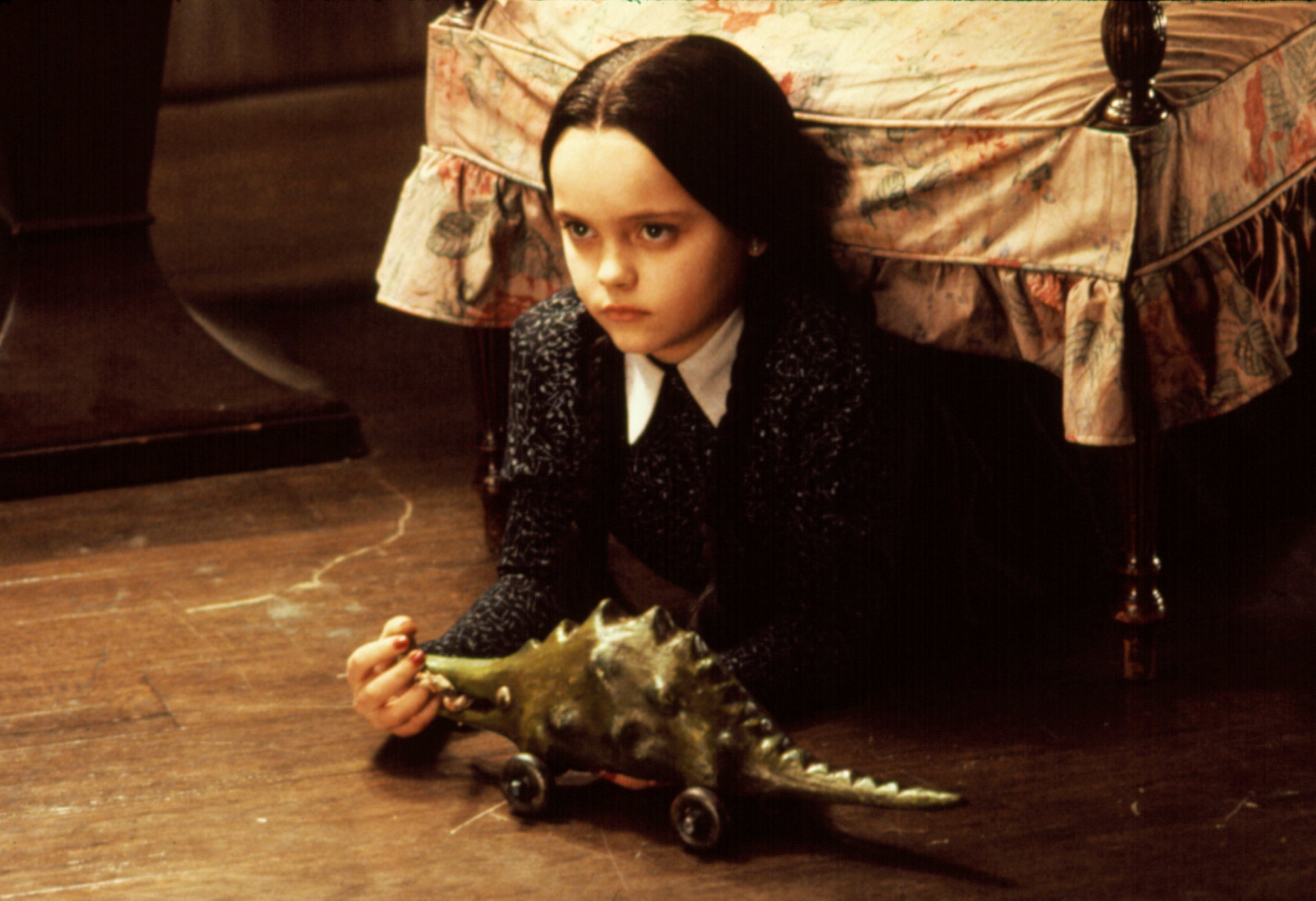 Christina Ricci as Wednesday Addams sits under a bed, holding a toy dinosaur on wheels, in a scene from the Addams Family movie