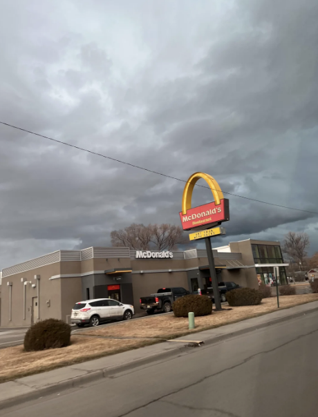 A McDonald's restaurant with a prominent golden arches sign on a cloudy day. Several vehicles are in the parking lot