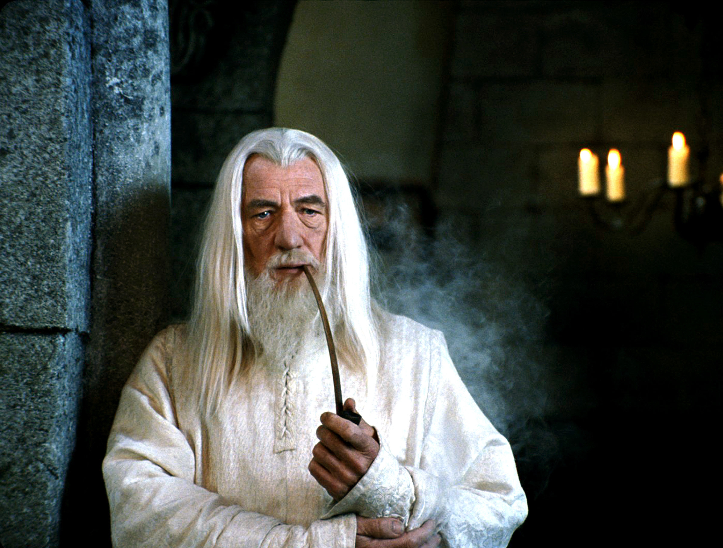 Gandalf from Lord of the Rings, played by Ian McKellen, stands indoors smoking a pipe, wearing a long white robe, with candles lit in the background