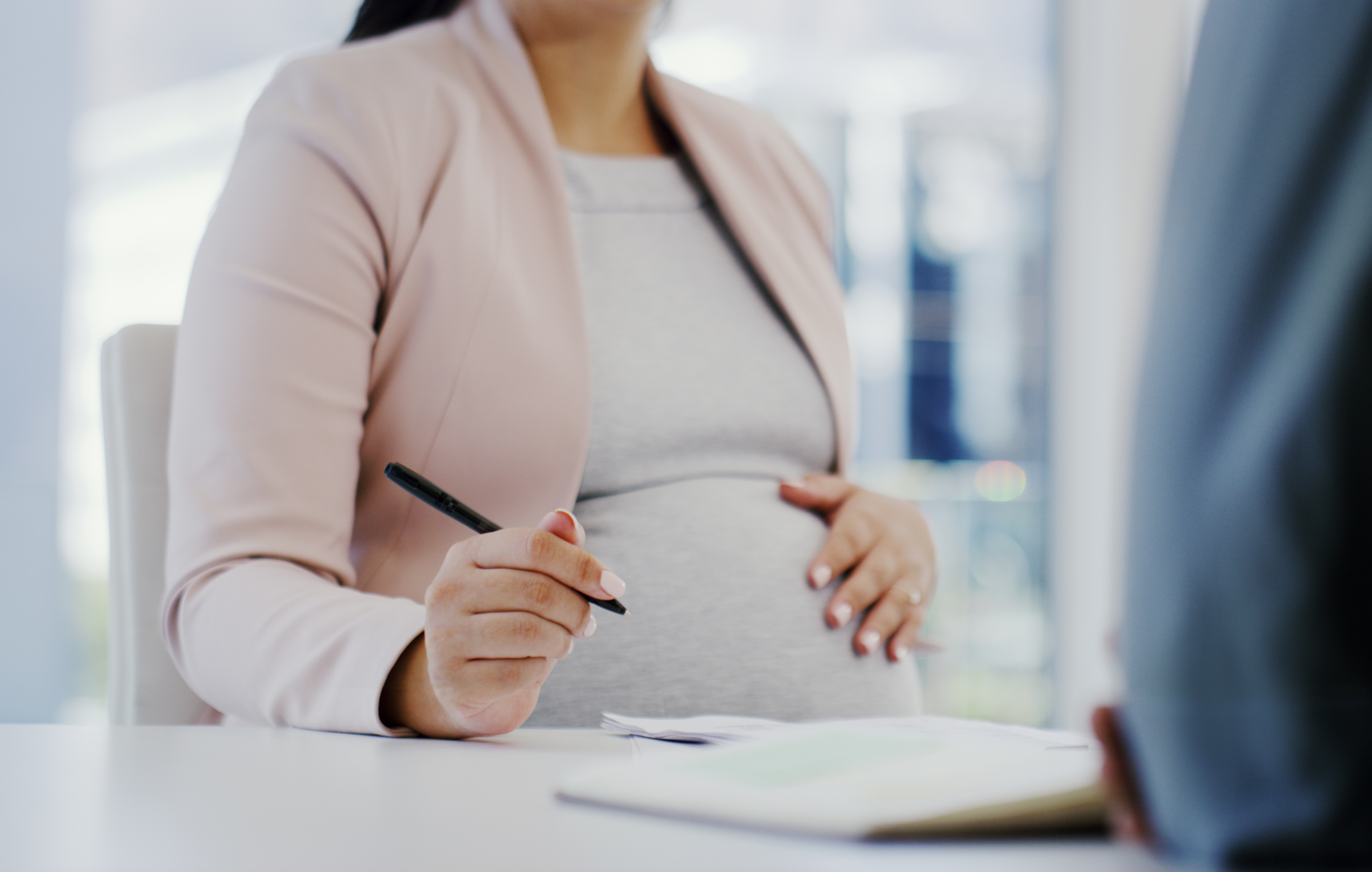 Pregnant woman in professional attire holds a pen and touches her belly, engaging in conversation at a desk with an unseen person