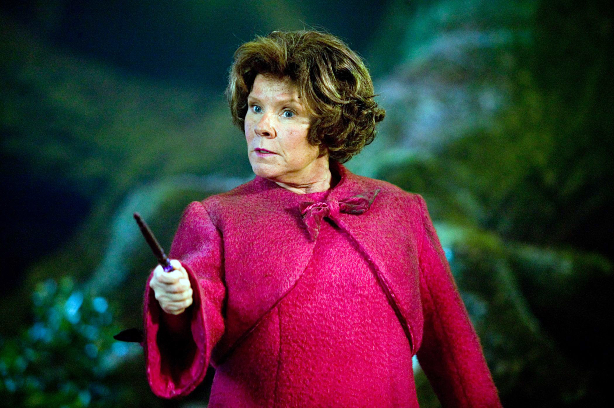 Imelda Staunton as Dolores Umbridge, dressed in a pink coat and holding a wand, in a forest scene from a movie