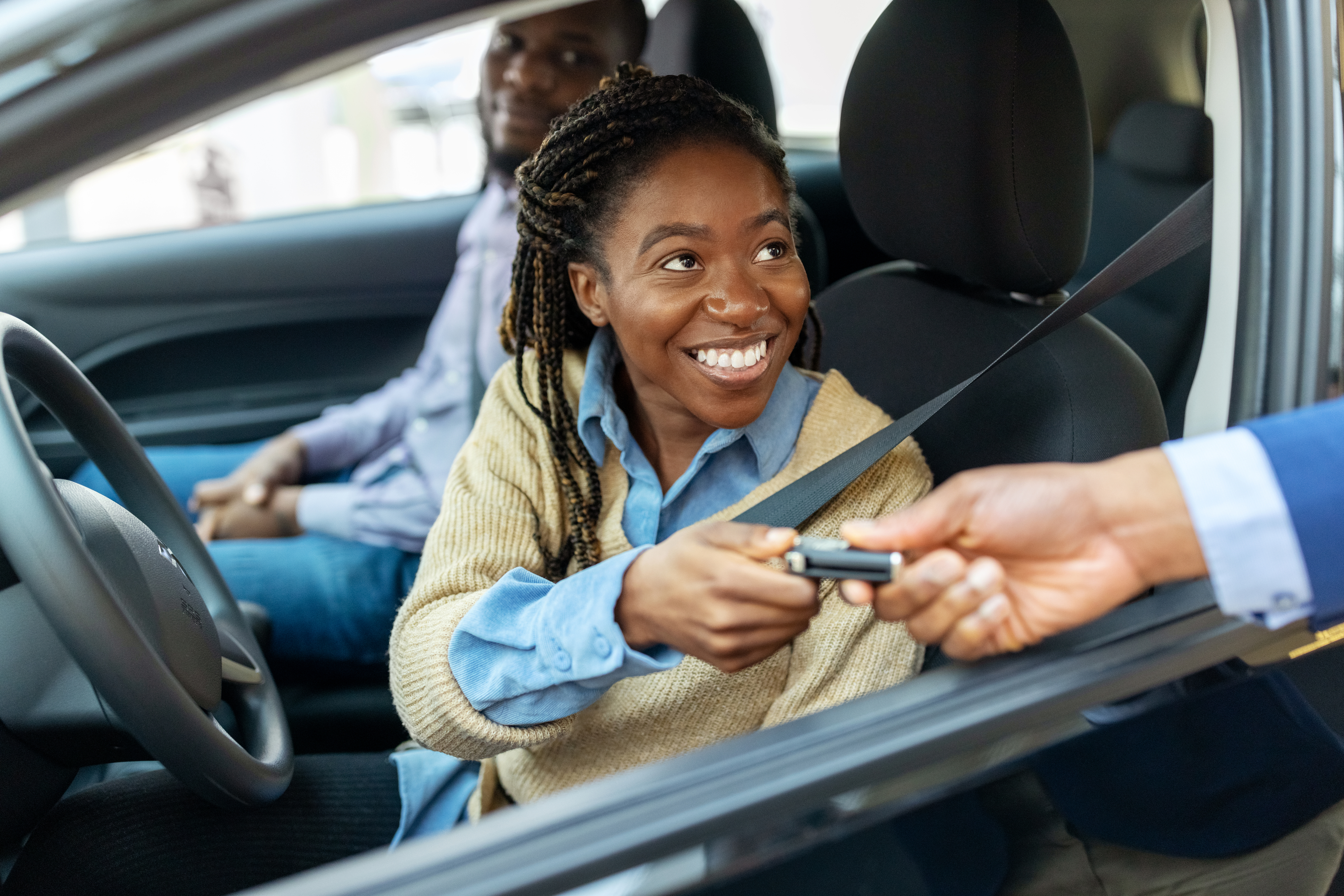 A smiling woman wearing a sweater and blue shirt receives car keys from a hand through the car window while a man sits in the passenger seat
