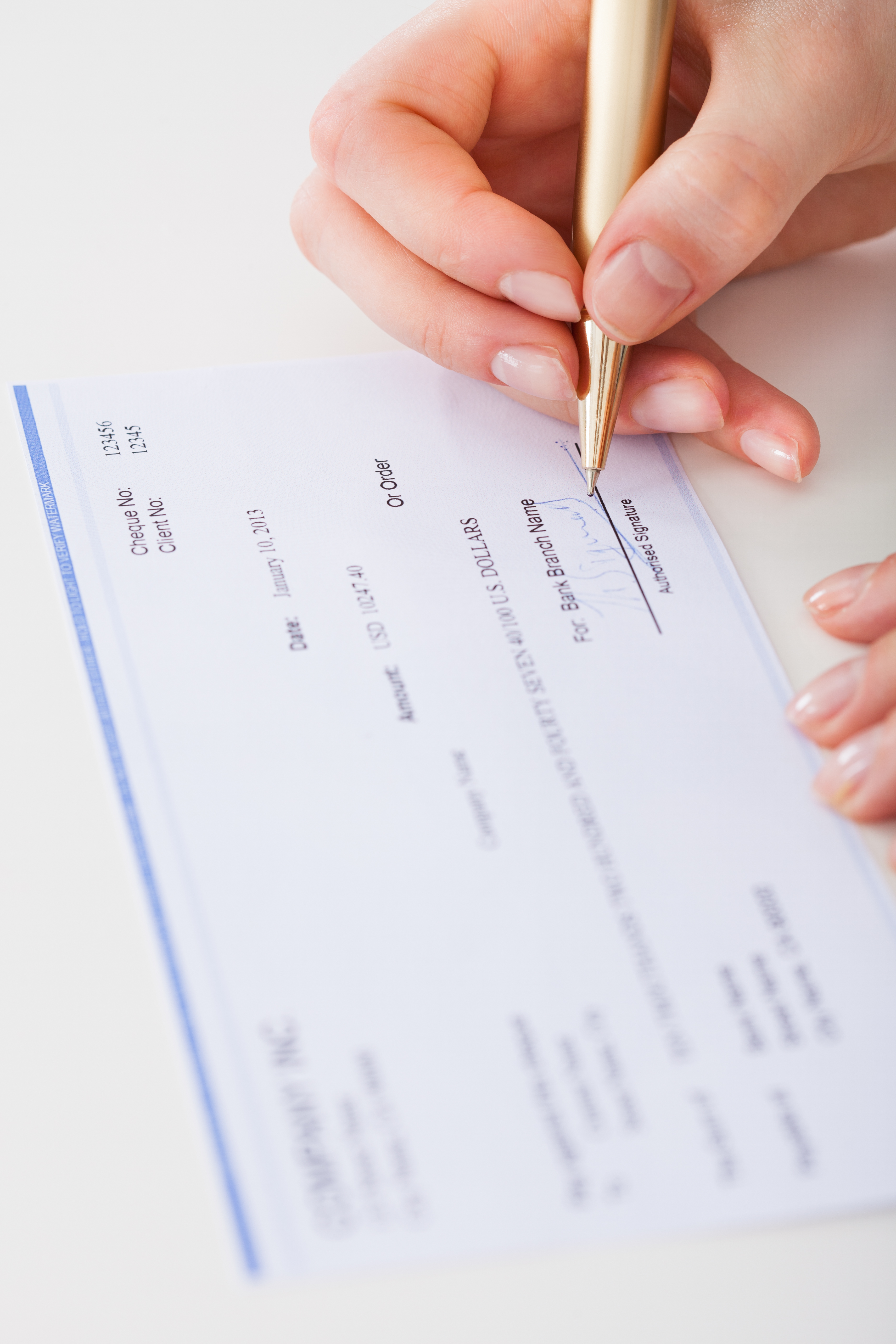 A close-up of a person signing a personal check with a gold pen. The check includes fields for amounts, dates, signatures, and recipient information