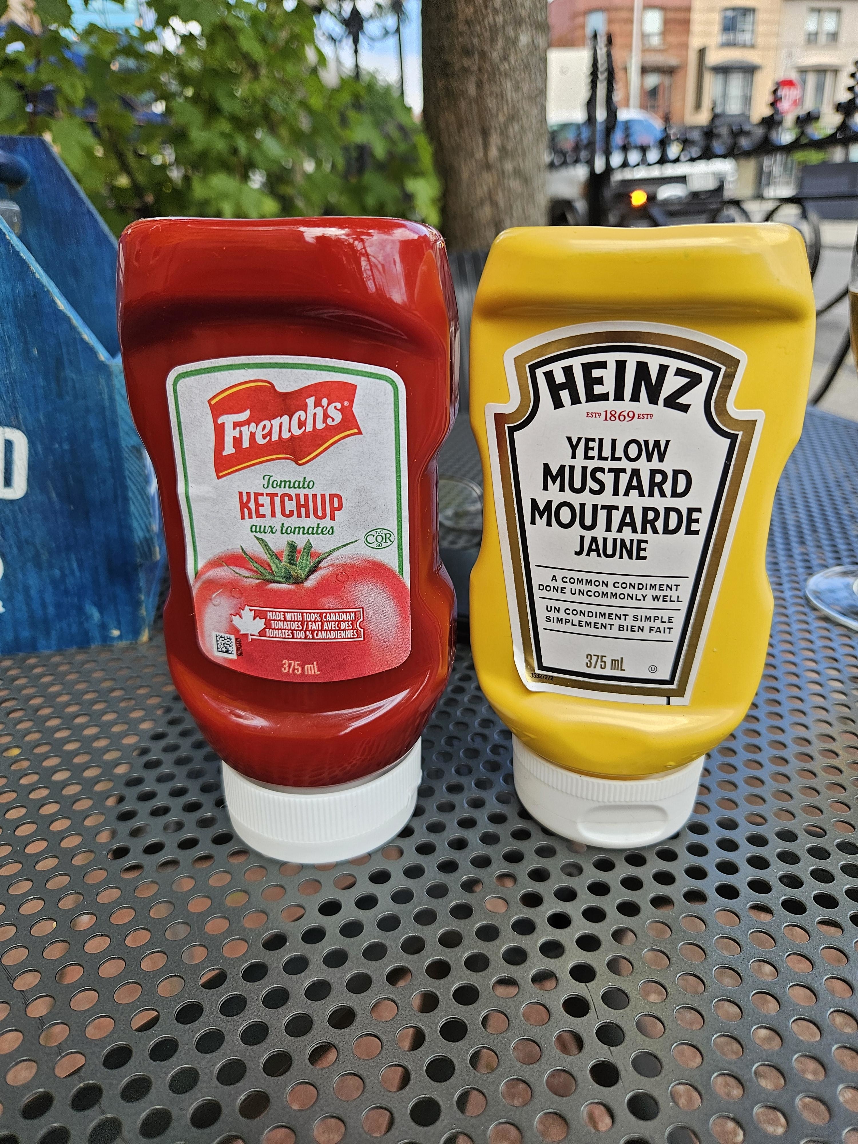 French's tomato ketchup bottle and Heinz yellow mustard bottle on a perforated metal table outdoors