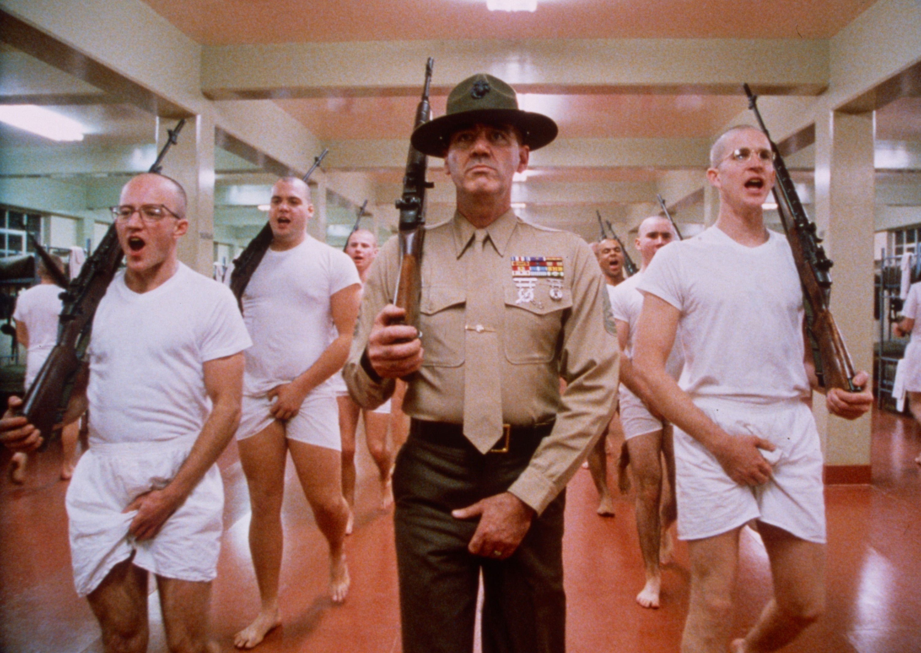 A scene from "Full Metal Jacket" with R. Lee Ermey as a drill instructor, leading soldiers in white undershirts and boxers during a training exercise, holding rifles