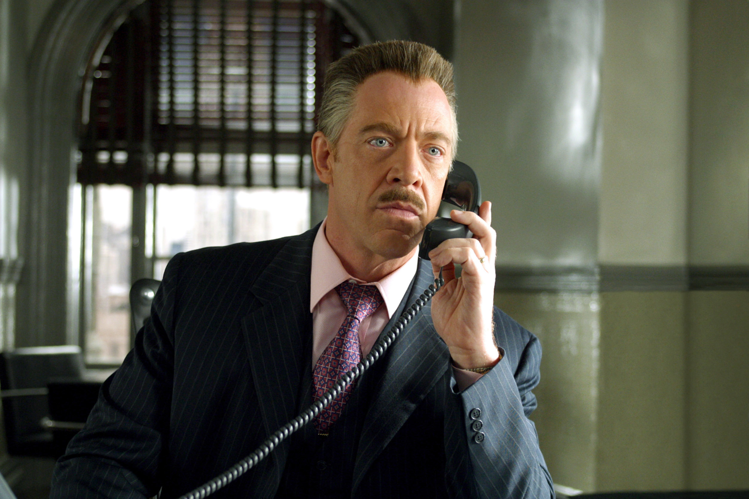 J.K. Simmons as J. Jonah Jameson, dressed in a pinstripe suit, holds a telephone while appearing serious in an office setting in a scene from a movie