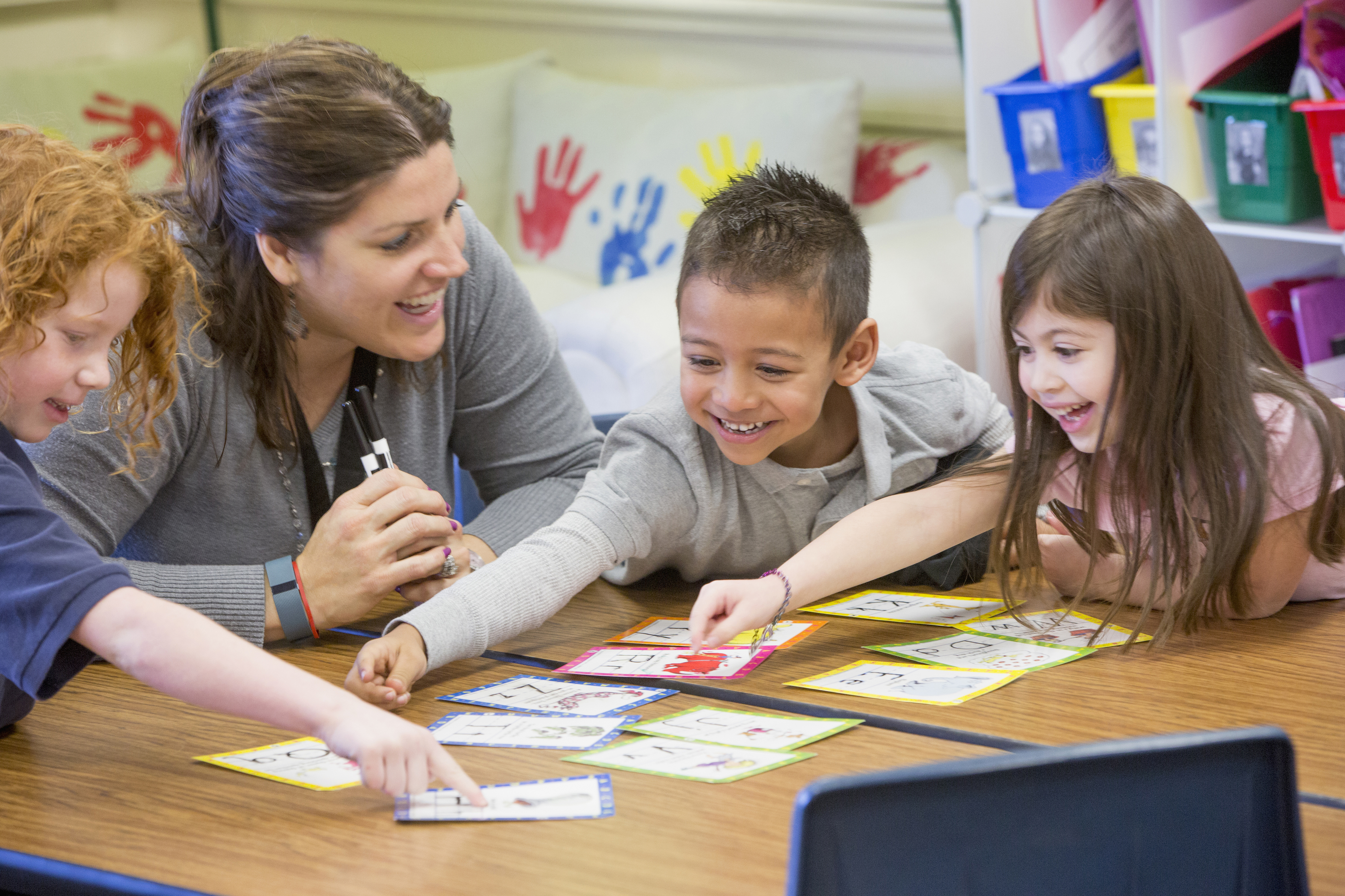 A teacher and three young children enthusiastically learn together at a table, pointing at educational cards