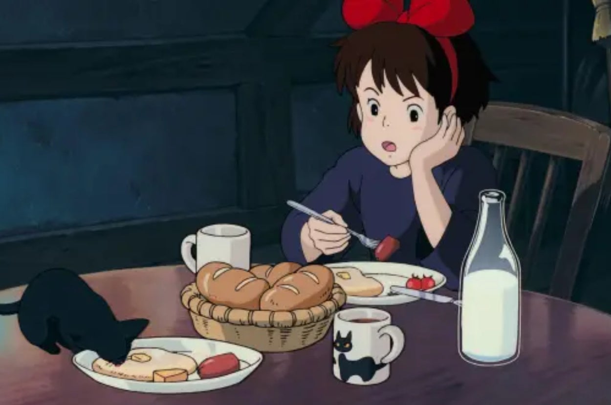 Kiki from Kiki's Delivery Service sits at a table with a black cat, Jiji, eating breakfast with bread, a fried egg, and milk
