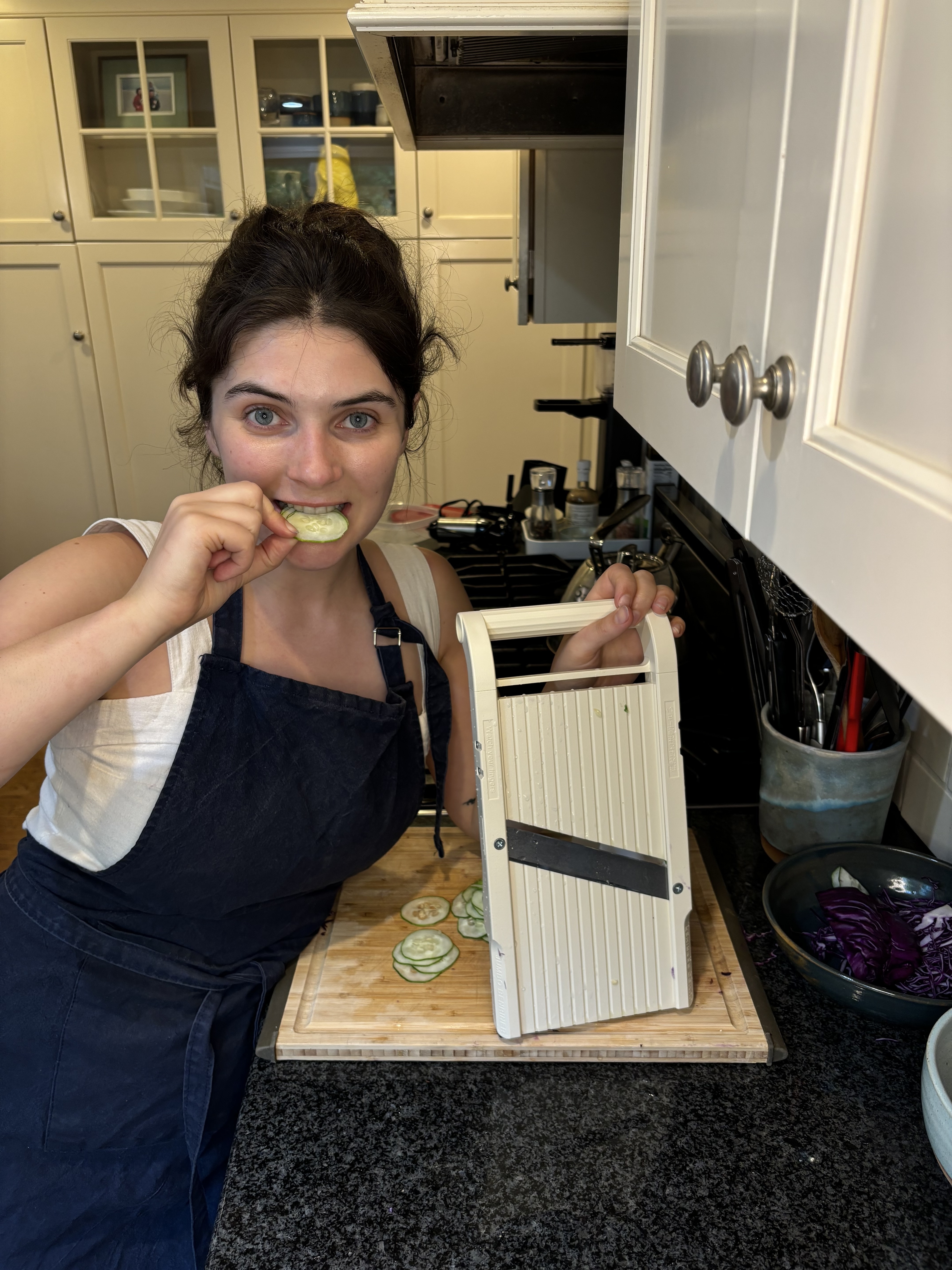 Woman in kitchen eating a cucumber slice, holding a mandoline slicer with sliced cucumbers on a cutting board
