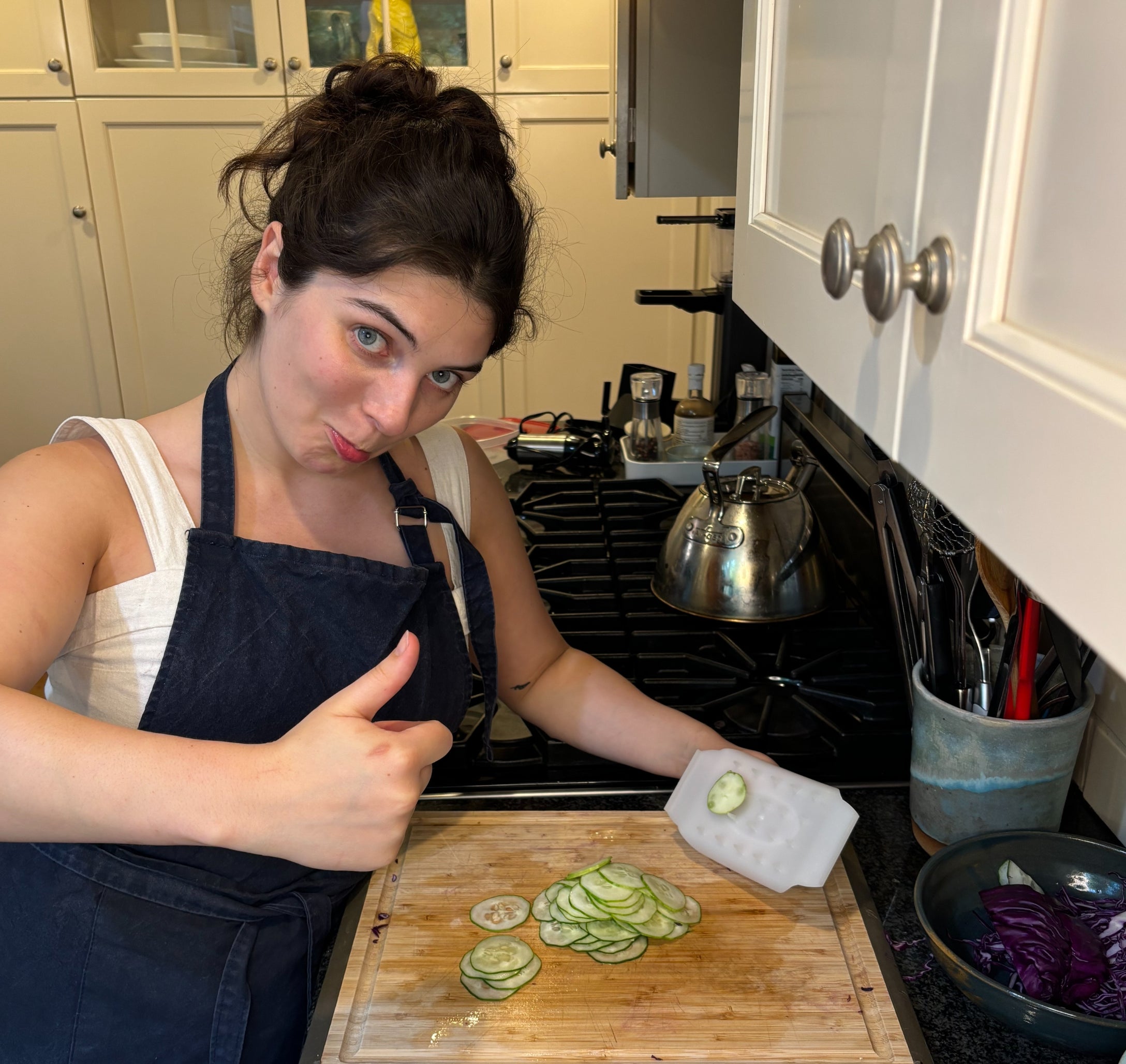 Person in a kitchen, wearing a blue apron and white top, gives a thumbs up while slicing cucumbers on a cutting board