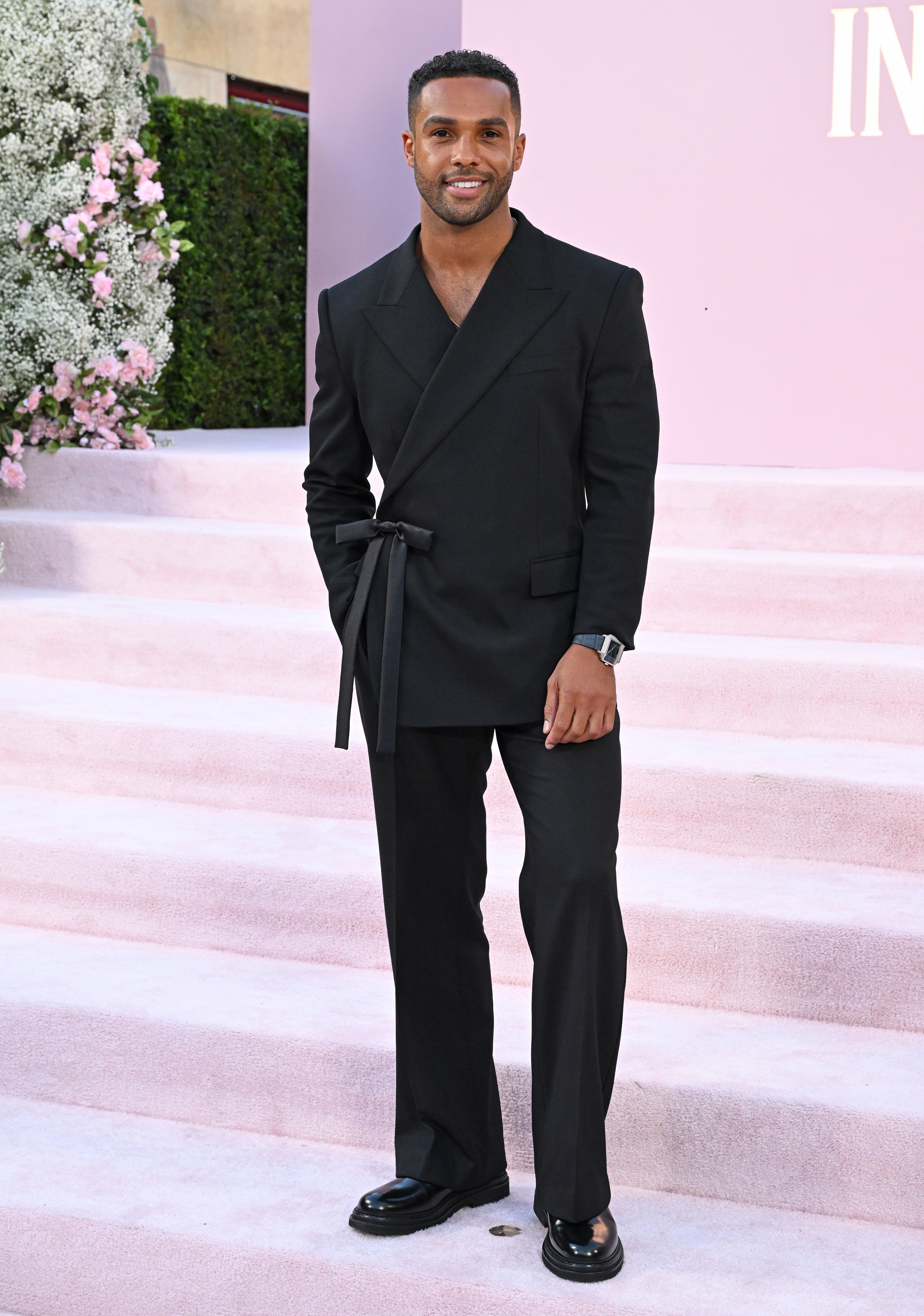 Aldis Hodge standing on a staircase in formal black attire with a belted blazer and dress shoes