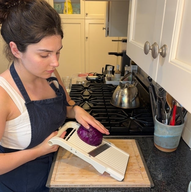 A person using a mandoline slicer to cut a purple cabbage in a kitchen. Various kitchen utensils are visible in the background