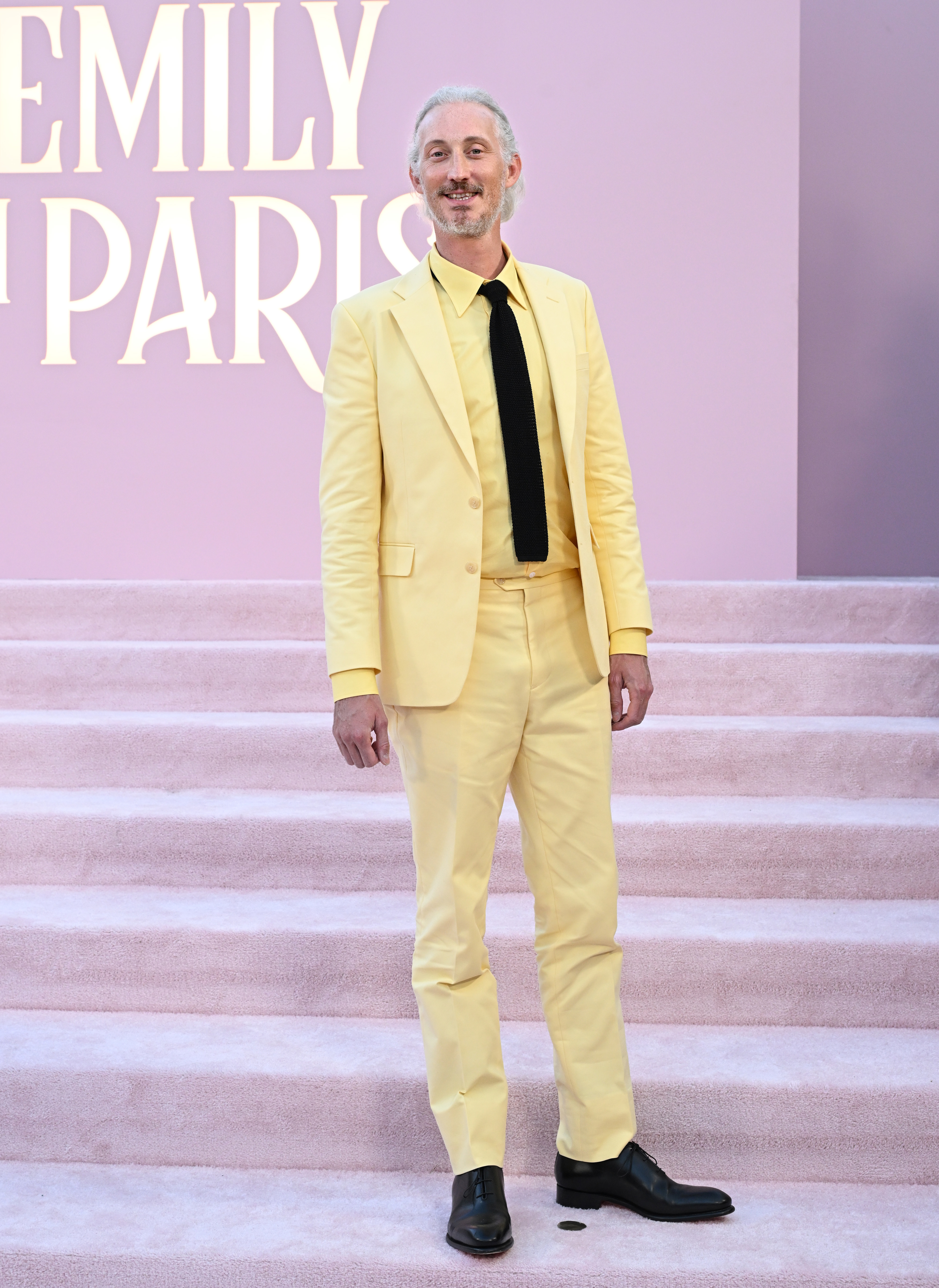 Man on a red carpet for "Emily in Paris," wearing a yellow suit with a black tie and black shoes