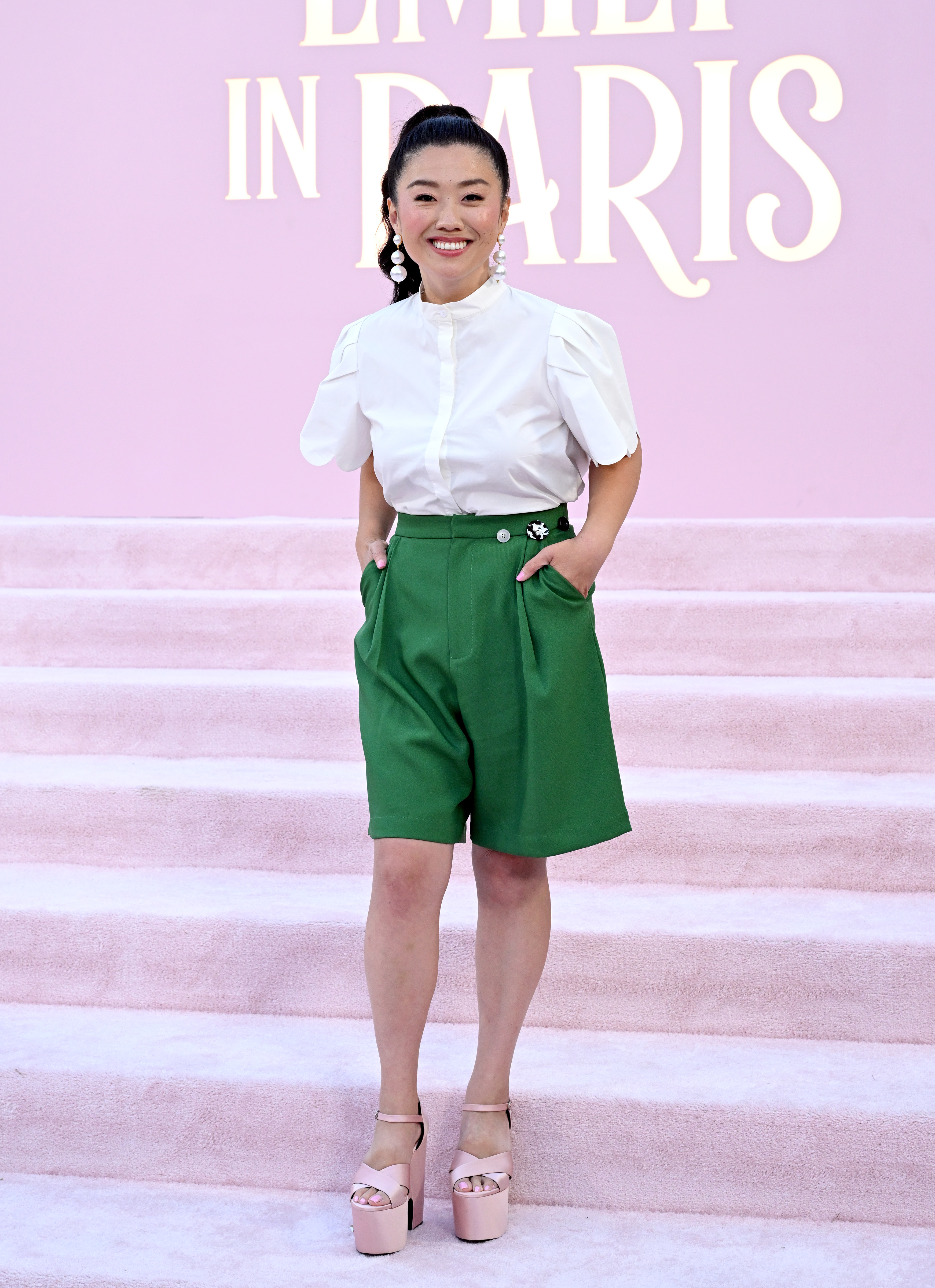 Sherry Cola stands on a light-colored carpet in front of an "Emily in Paris" sign, wearing a white blouse, green high-waisted shorts, and platform heels