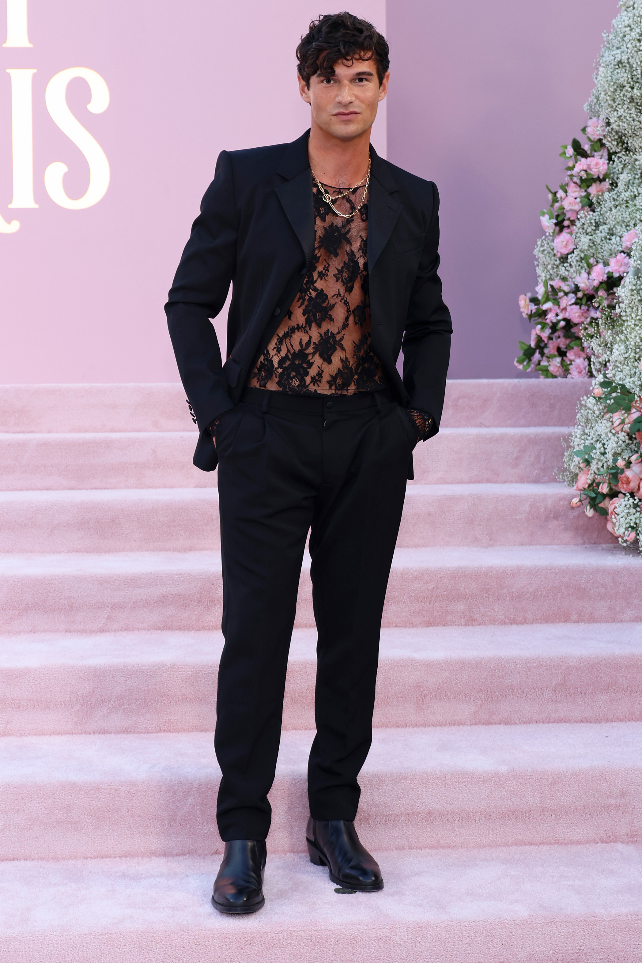 Man in a black suit with a see-through lace shirt standing on pink carpet by floral arrangement
