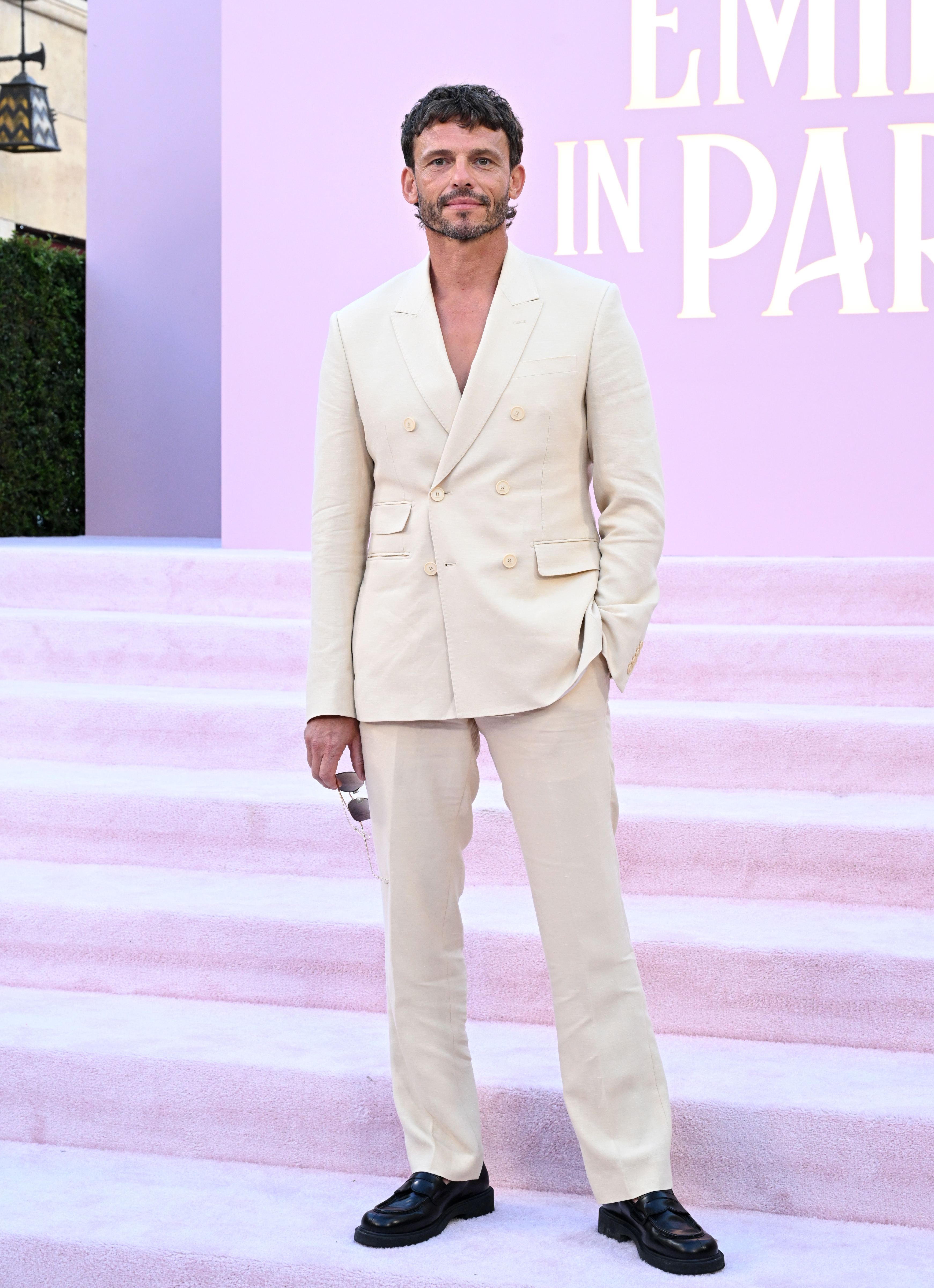 Arnaud Binard stands on pink steps in a light-colored suit with black loafers at the "Emily in Paris" event