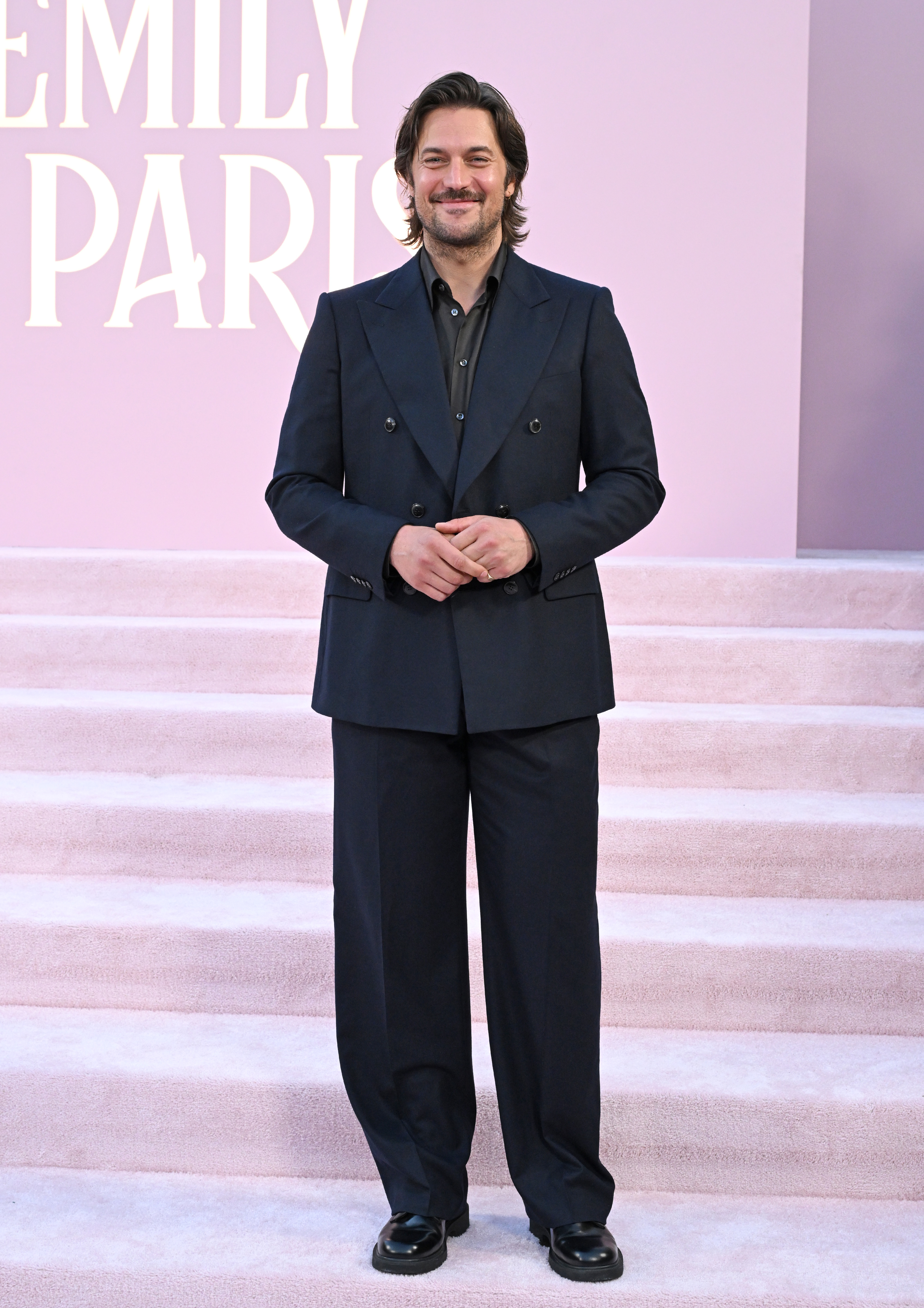 Lucas Bravo stands on steps in a dark double-breasted suit, smiling at the camera. The text "Emily in Paris" is partially visible in the background