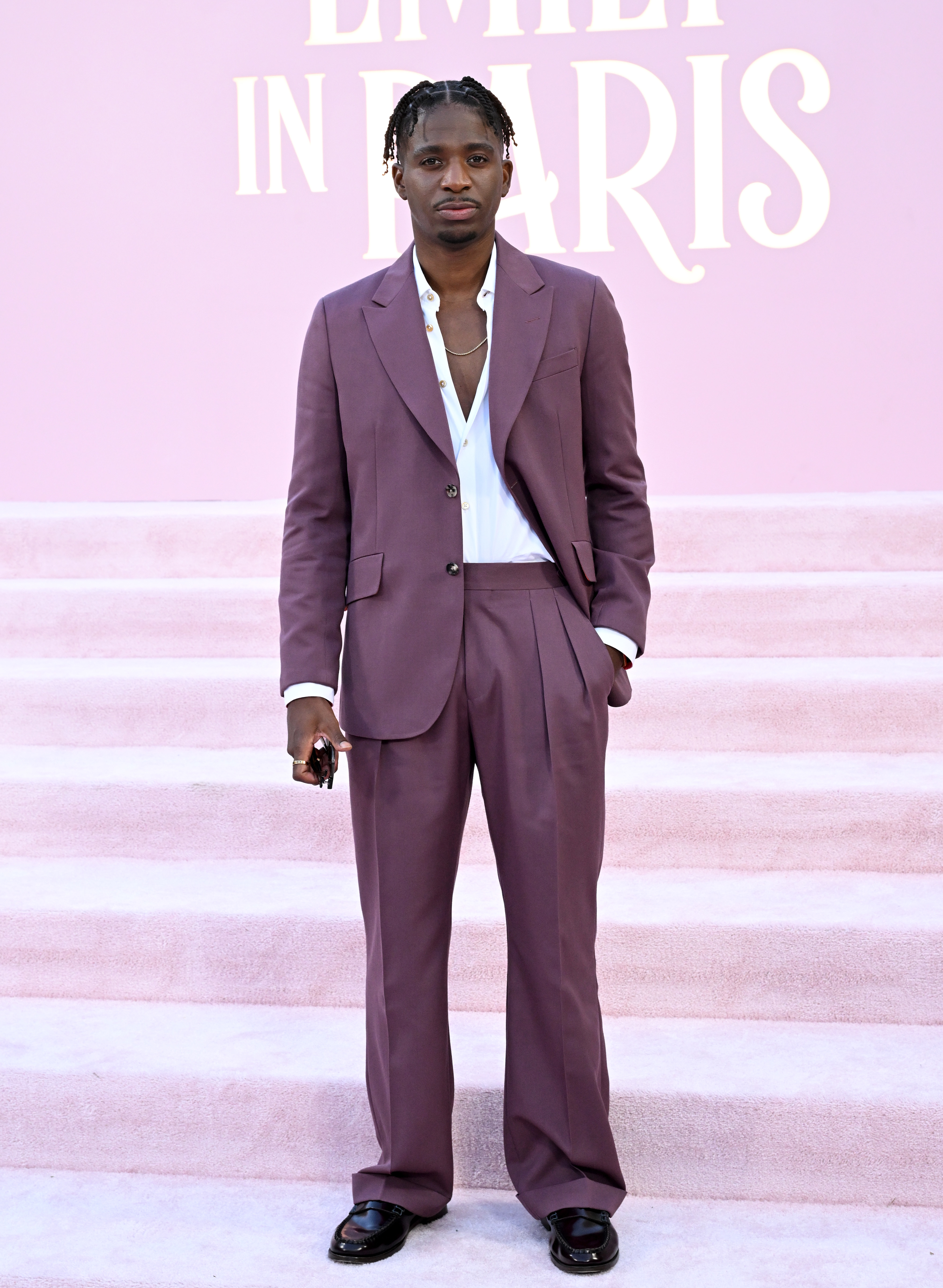 Lucien Laviscount is on a pink carpet, wearing a mauve suit with a white shirt and black shoes. He is holding a phone in his right hand