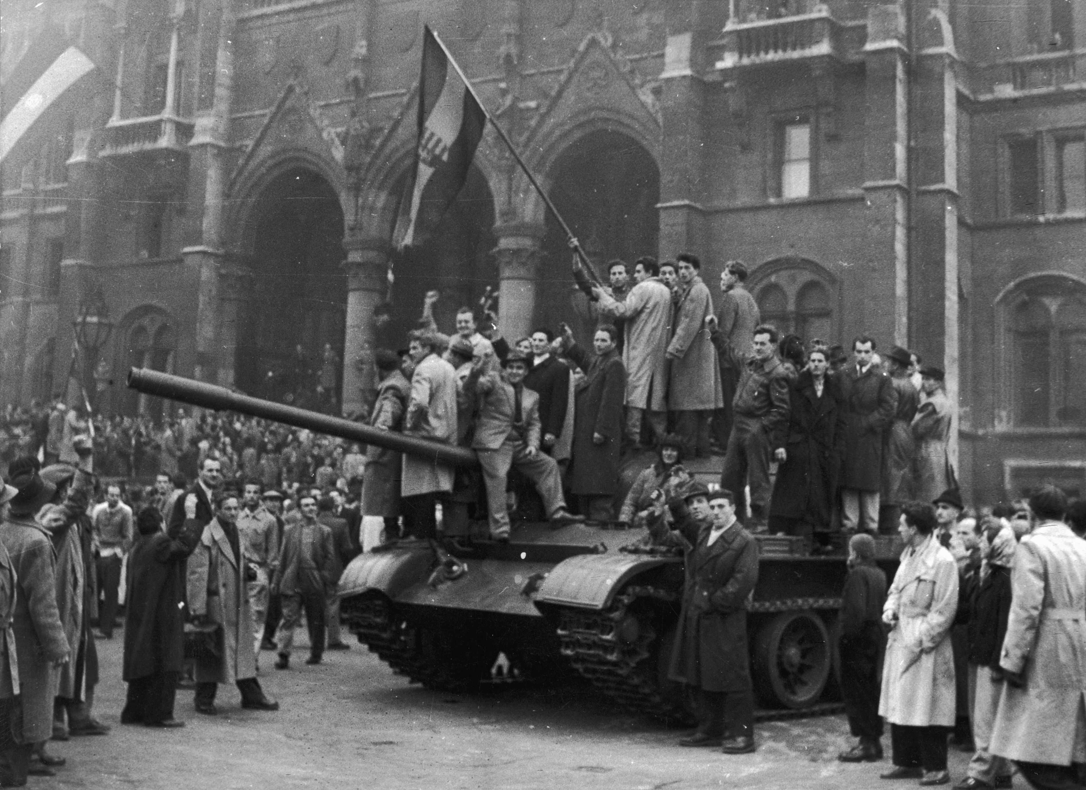 People gathered around and on top of a tank in front of a large building during a historical event, waving a flag and cheering