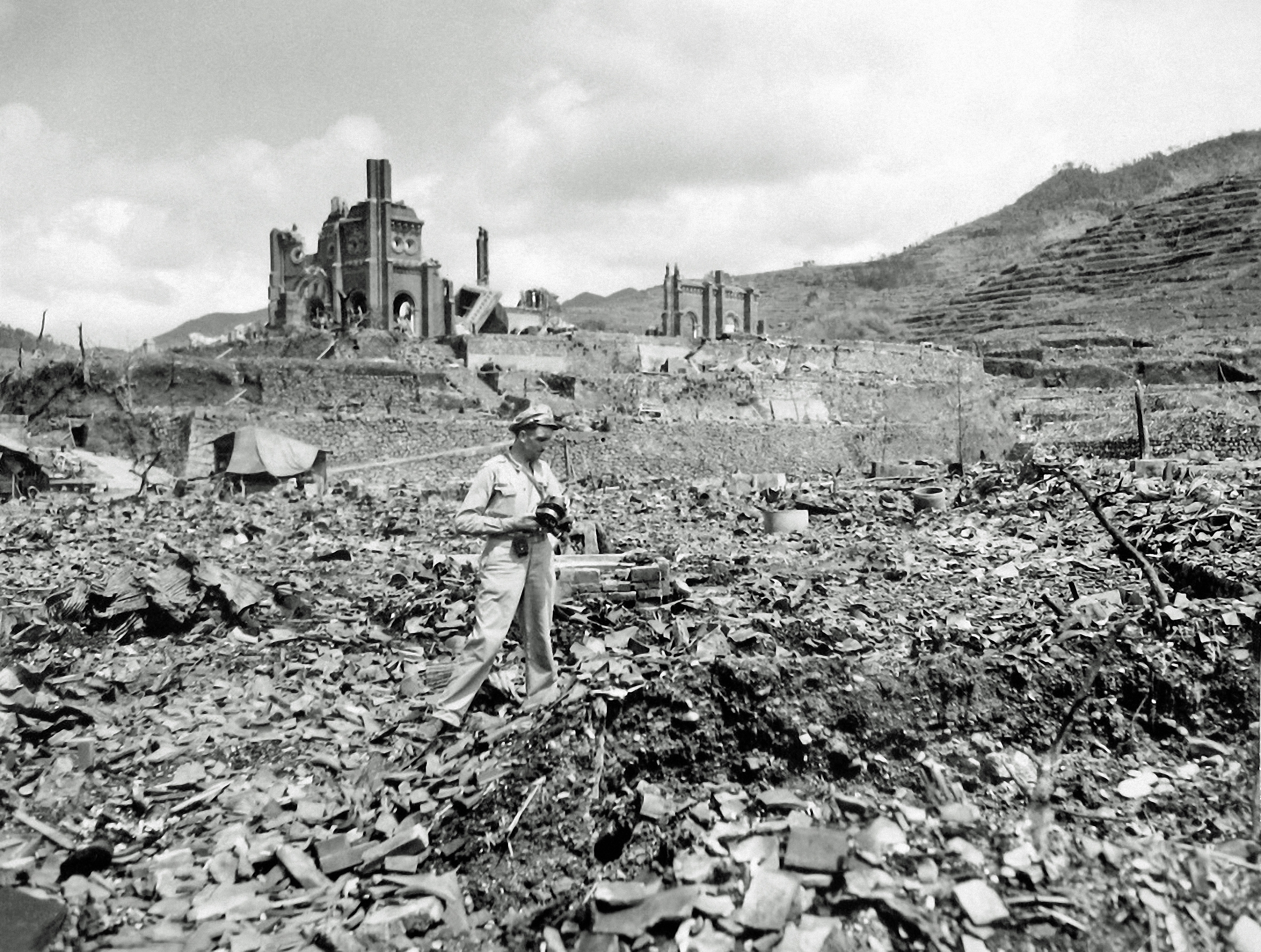 A man in military uniform walks through the ruins and rubble of buildings after a bombing, with a partially destroyed structure in the background