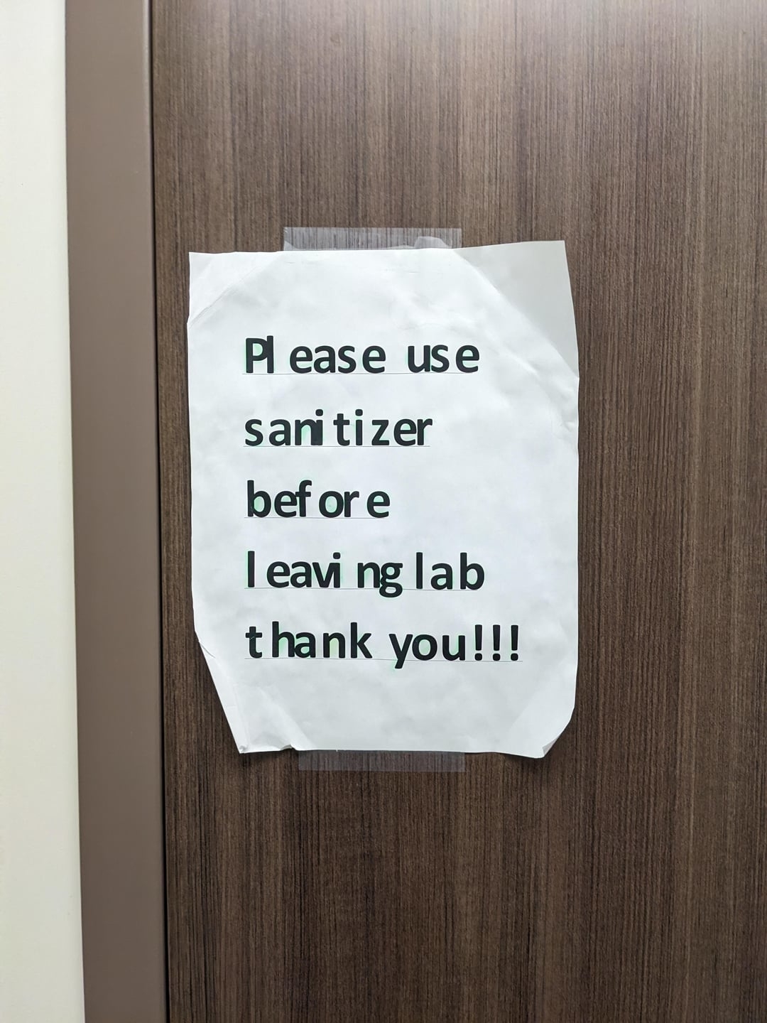 Sign taped to a wooden door reads: "Please use sanitizer before leaving lab thank you!!!" but the spaces in between the letters are all differently sized