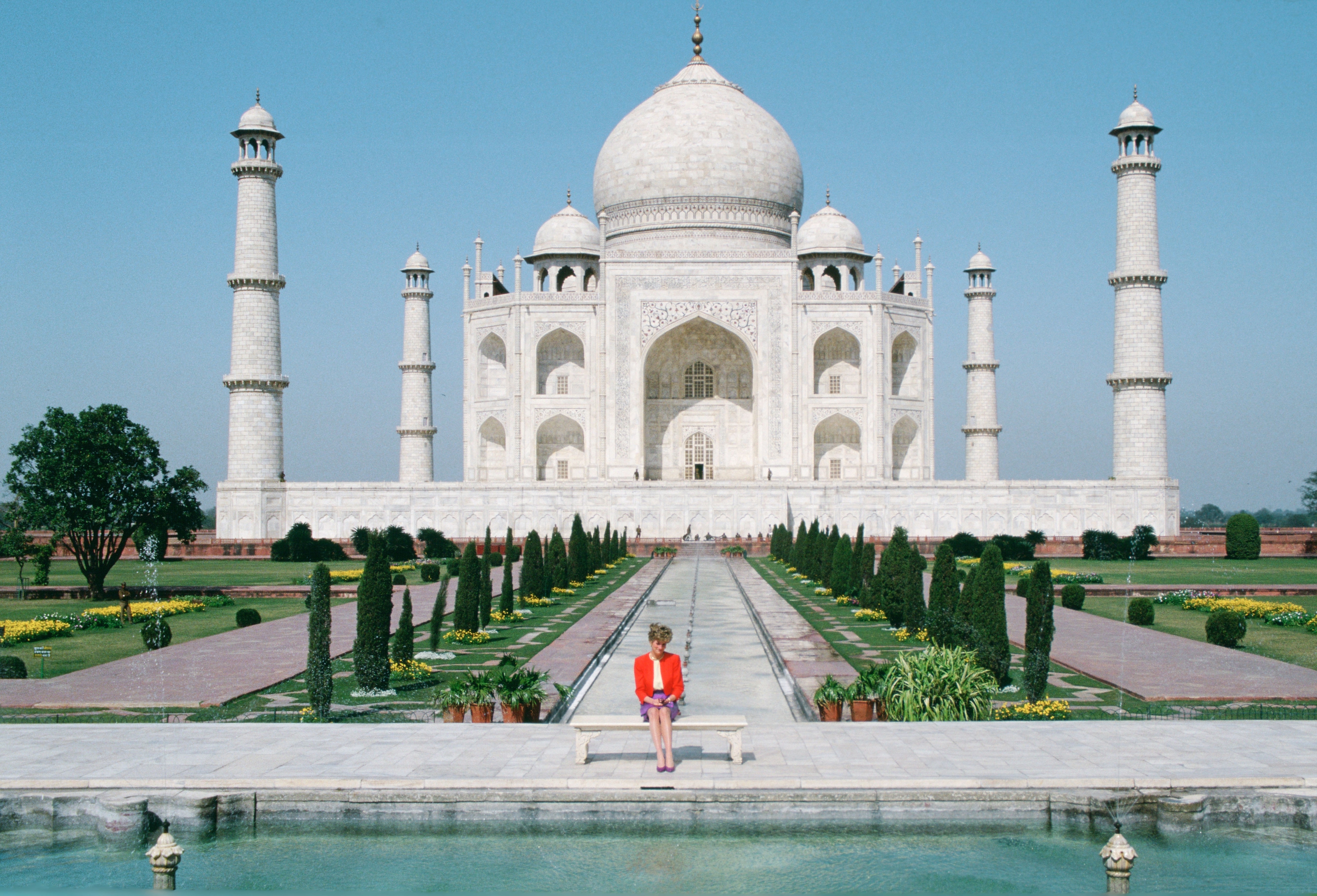 A person sits on a bench facing the Taj Mahal, a large white marble mausoleum with four minarets, lush gardens, and a reflecting pool in the foreground