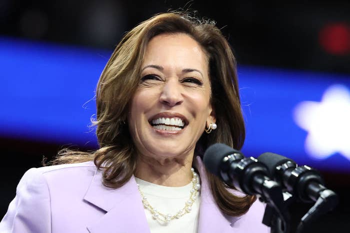Kamala Harris smiles at a podium wearing a light-colored blazer and pearl necklace. A blurred blue background is behind her
